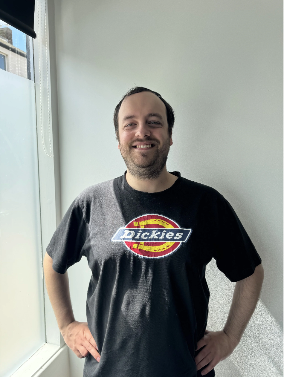 A person with short black hair, wearing a Dickies t-shirt, standing in front of a white wall.
