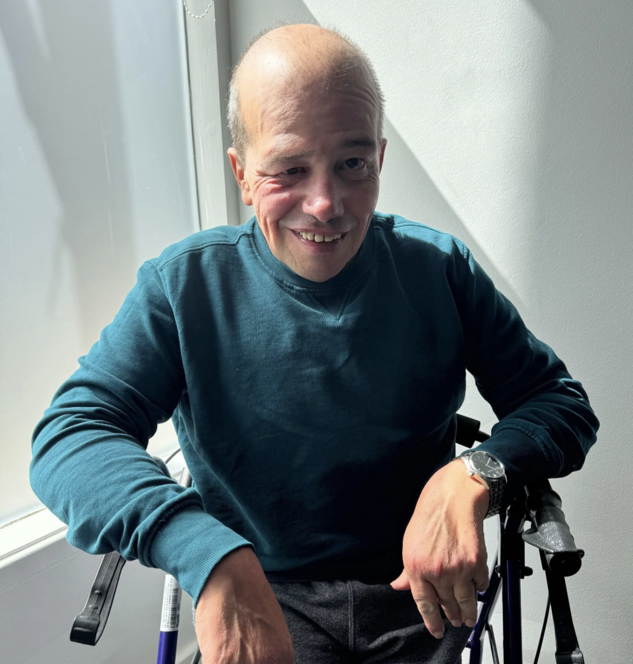 Smiling person in a blue shirt, sat on a walker in front of a white wall.