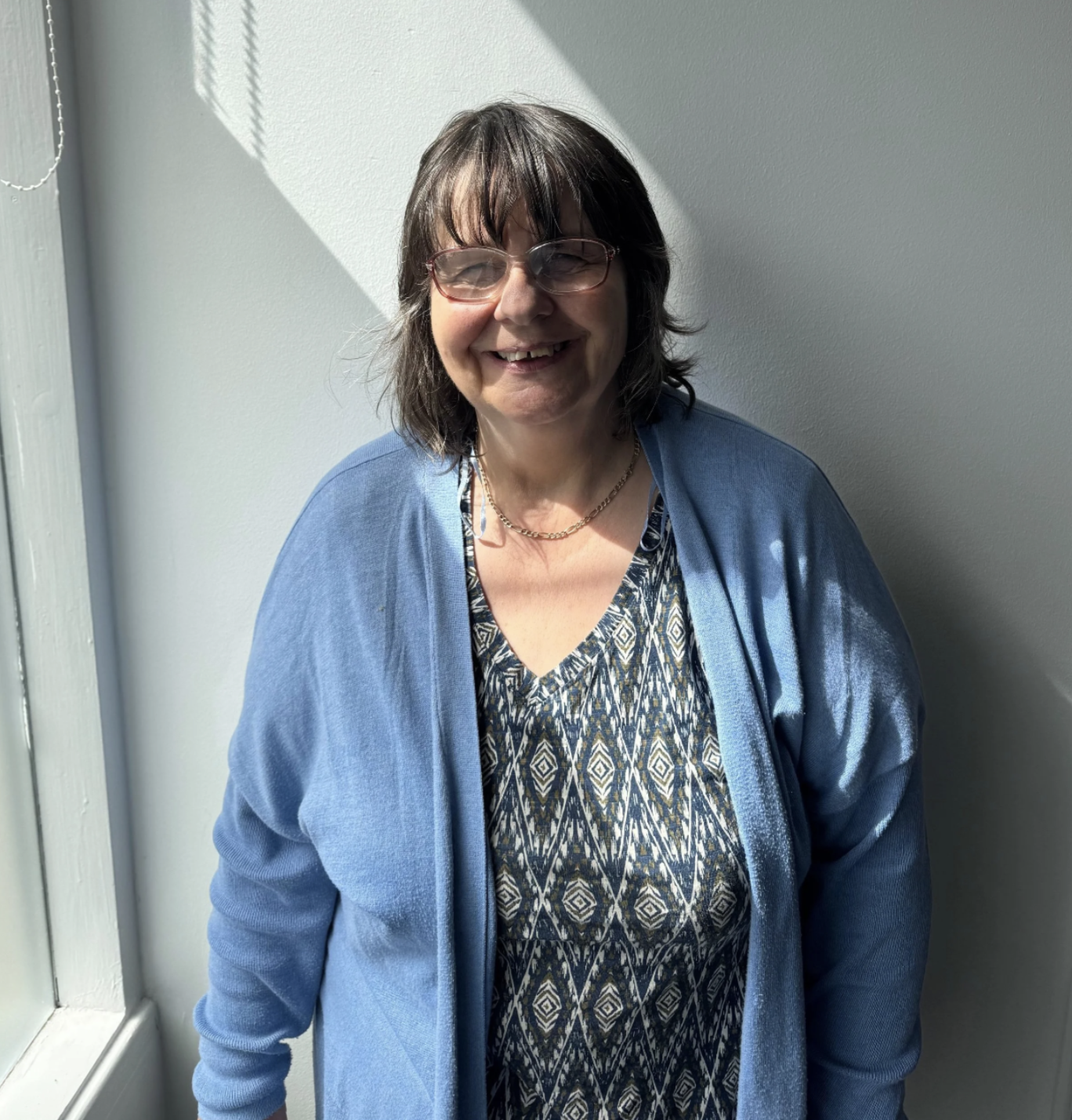 A person with short, dark hair, wearing glasses, a blue cardigan and a grey, diamond print dress. She is standing in front of a white wall.