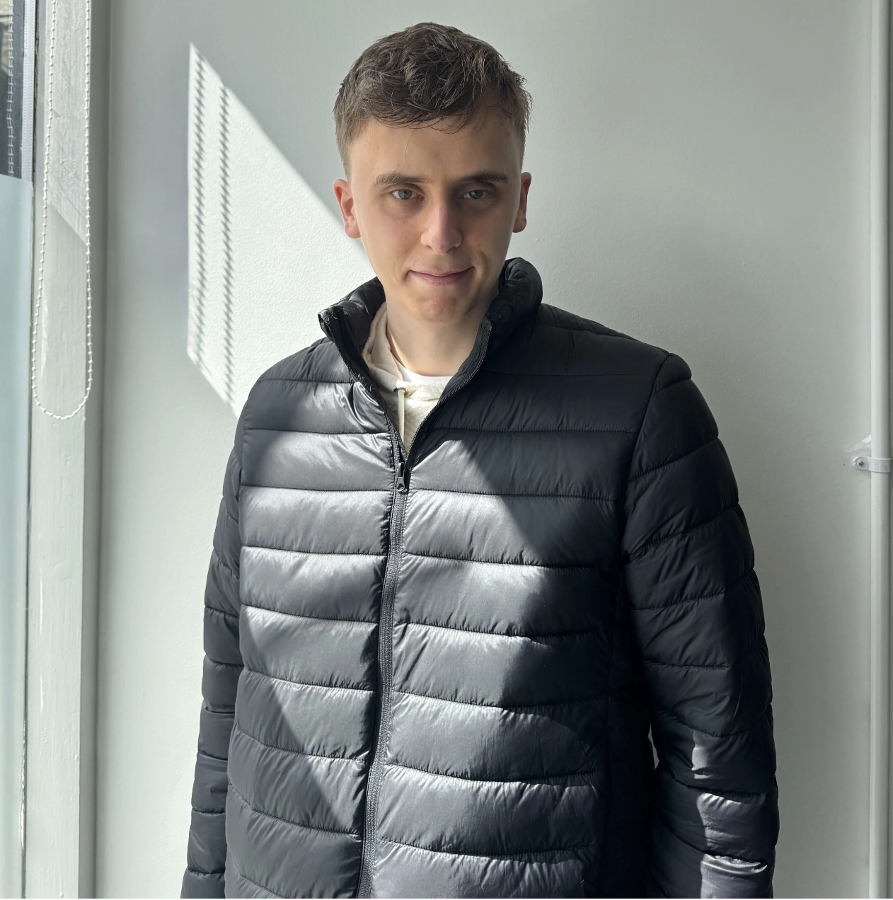 A man with short brown hair, wearing a puffer jacket, standing in front of a white wall.