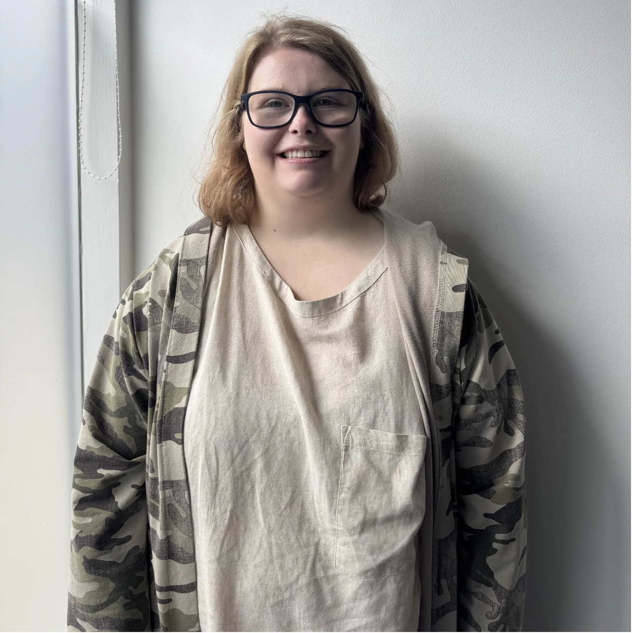 A person with short blond, brown hair, wearing glasses, a light brown t-shirt and a camouflage jacket. They are standing in front of a white wall.