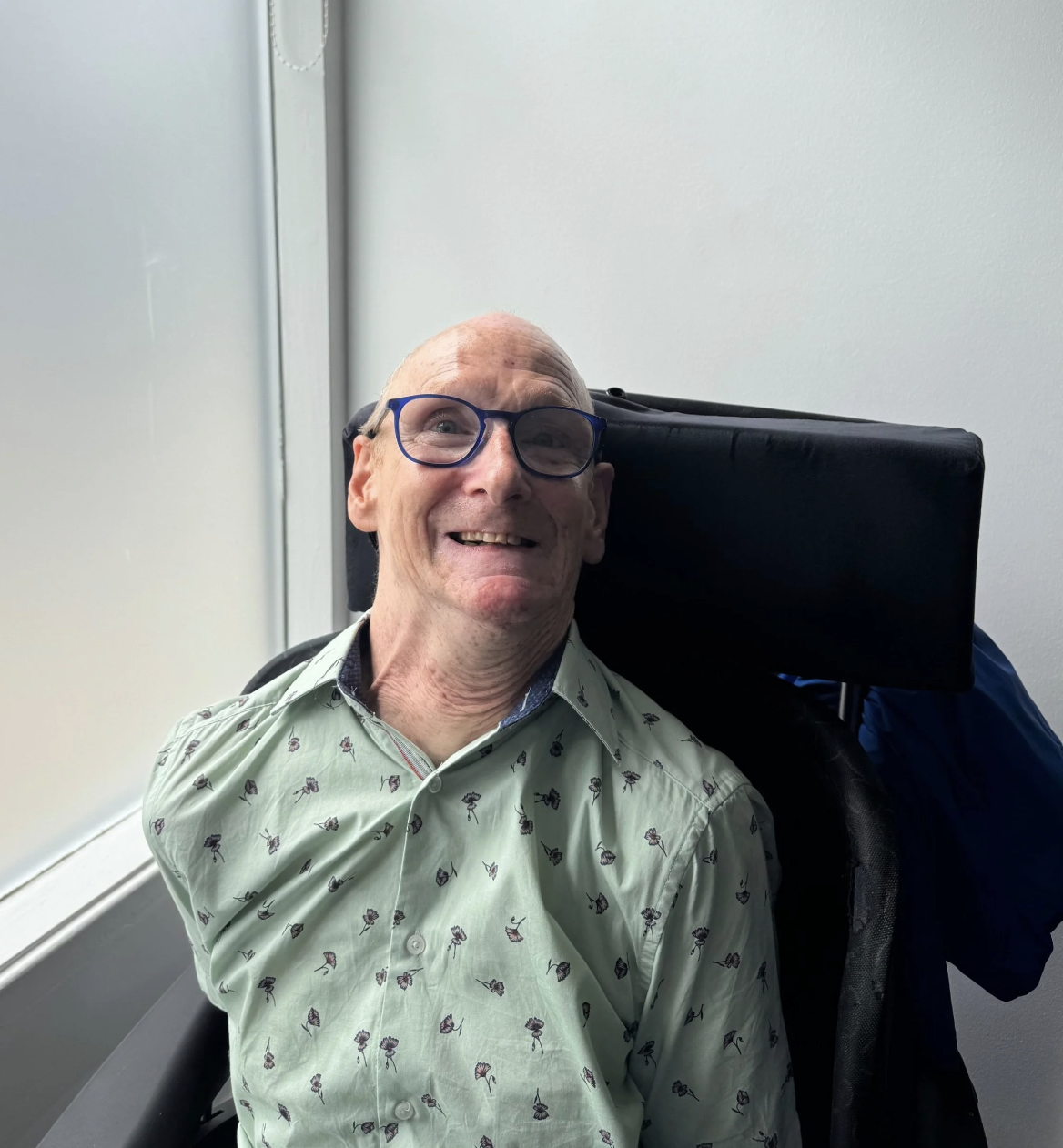 A bald person in a wheelchair with glasses, wearing a green flowery top.