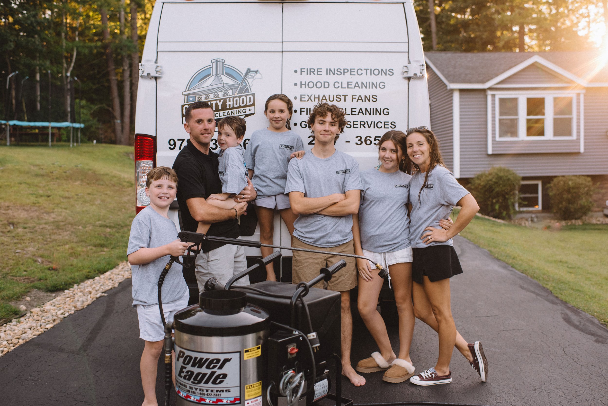 A group of six people, five young adults and one child, standing in front of a cleaning service van during sunset. One person holds a power washer, and the van displays company services for fire inspections, hood cleaning, exhaust fans, and fire cleaning.