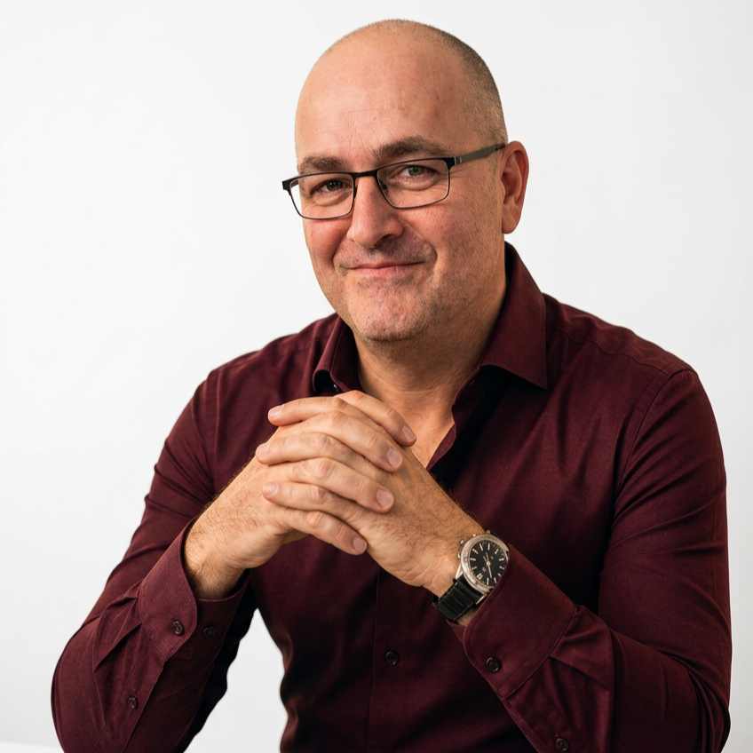 Un homme avec des lunettes portant une chemise bordeaux, souriant, assis avec ses mains jointes contre un fond blanc.