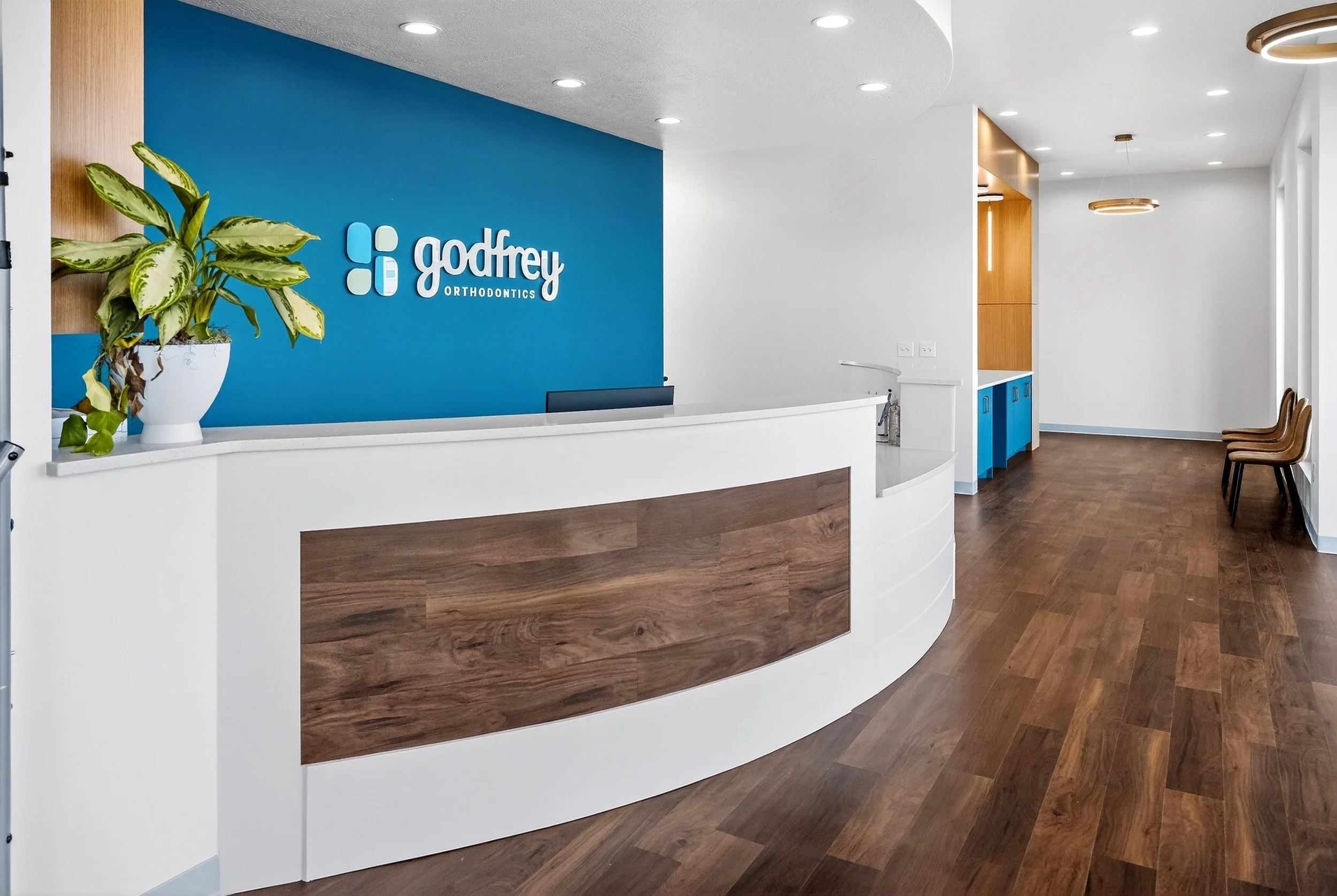 Modern orthodontic clinic reception area with white desk and blue accent wall displaying the 'Godfrey Orthodontics' logo, wooden flooring, and seating area along the wall.