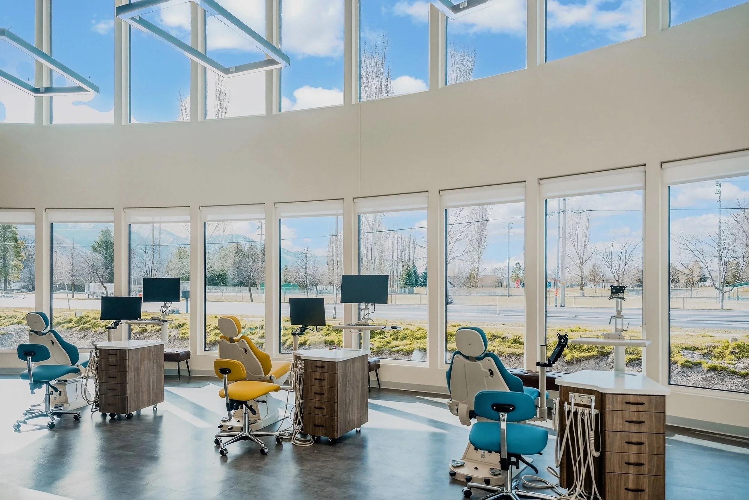 Empty modern Orthodontic clinic with dental chairs, monitors, and large windows showing a view of trees and a blue sky.