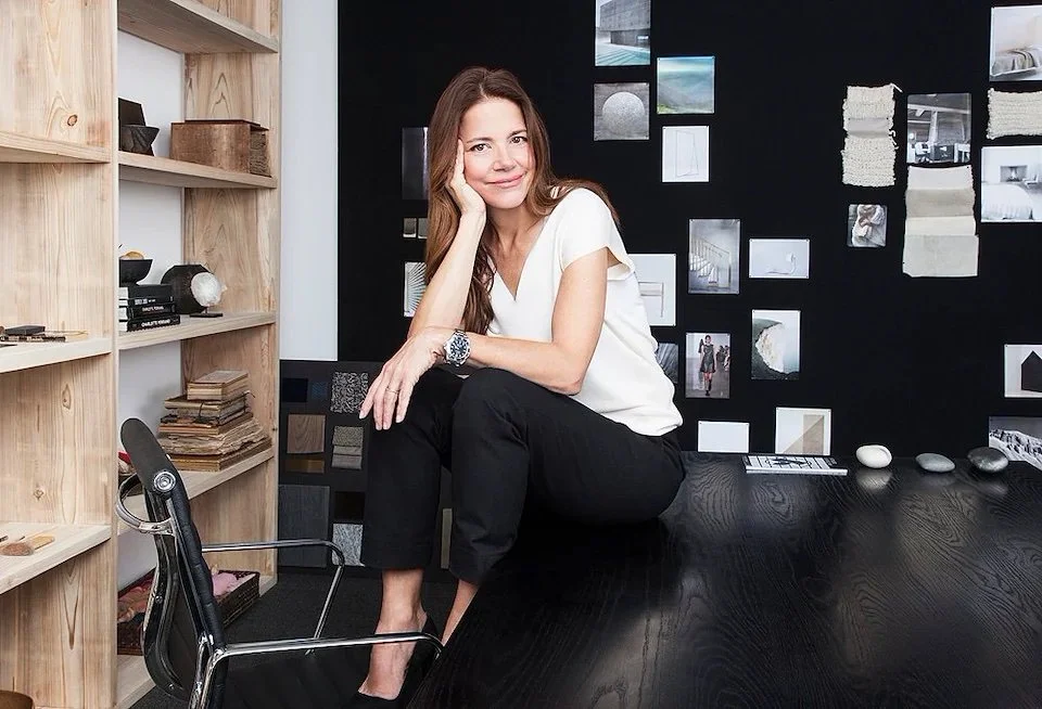 A woman with long brown hair sitting on a black desk in a modern office or art studio, smiling and resting her head on her hand. She has beautiful, natural, easy makeup and hair by Maysoon Faraj in NYC