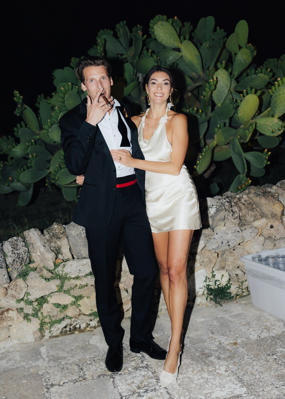 A man and a woman dressed in formal attire standing outdoors at night in front of large green cactus plants. The man is smoking a cigar and wearing a black tuxedo with a white shirt, while the woman is smiling and wearing a sleeveless satin dress wit