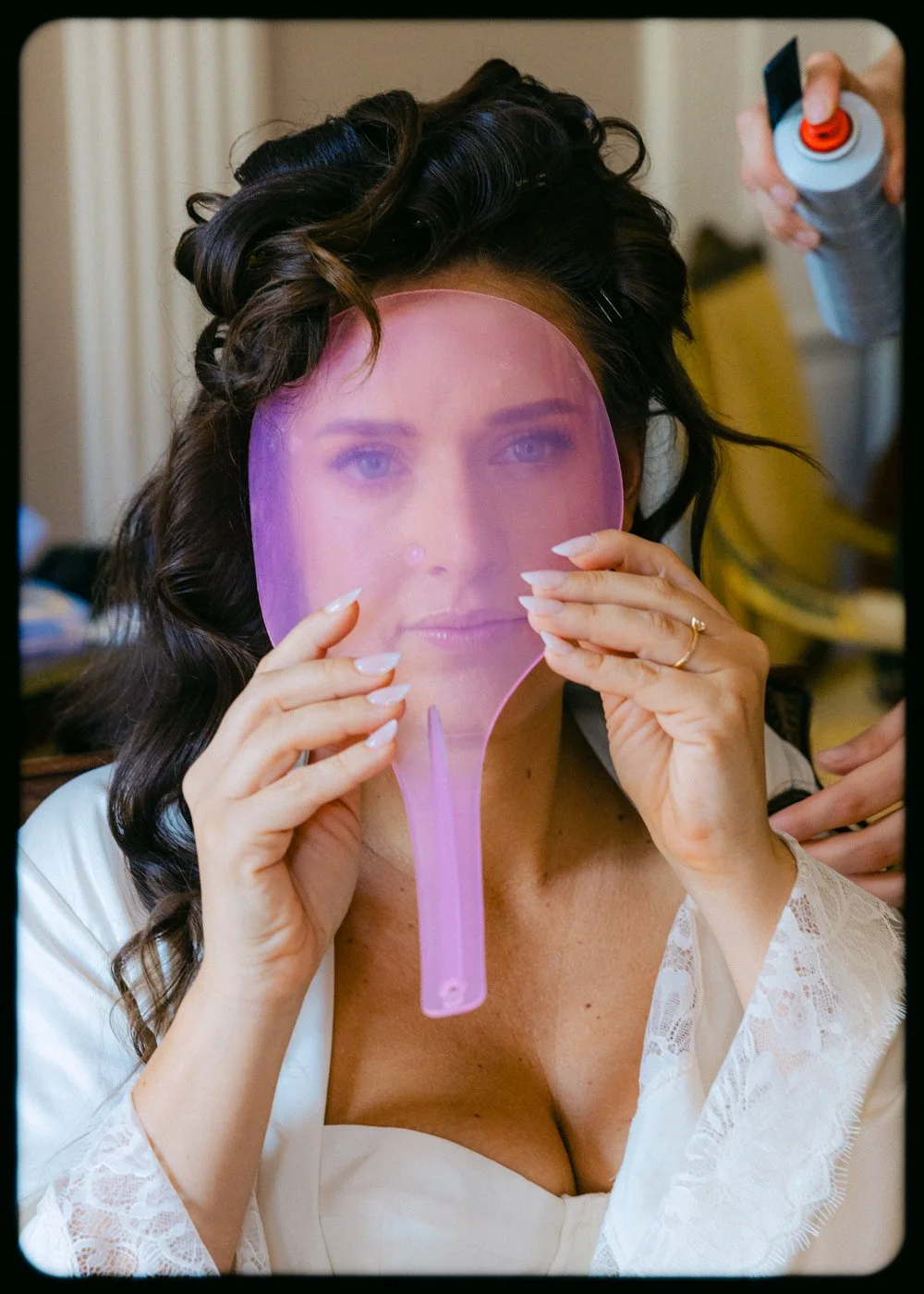 Woman with dark, curly hair holding a pink handheld mirror in front of her face, looking into it, while someone sprays her hair with a can of hairspray in the background.