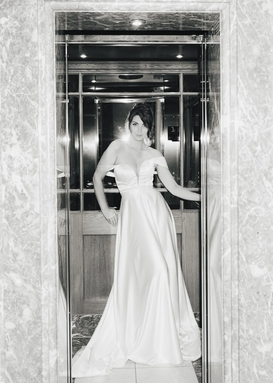 Elegant bride posing confidently in a satin gown inside a mirrored elevator, captured in timeless black and white.