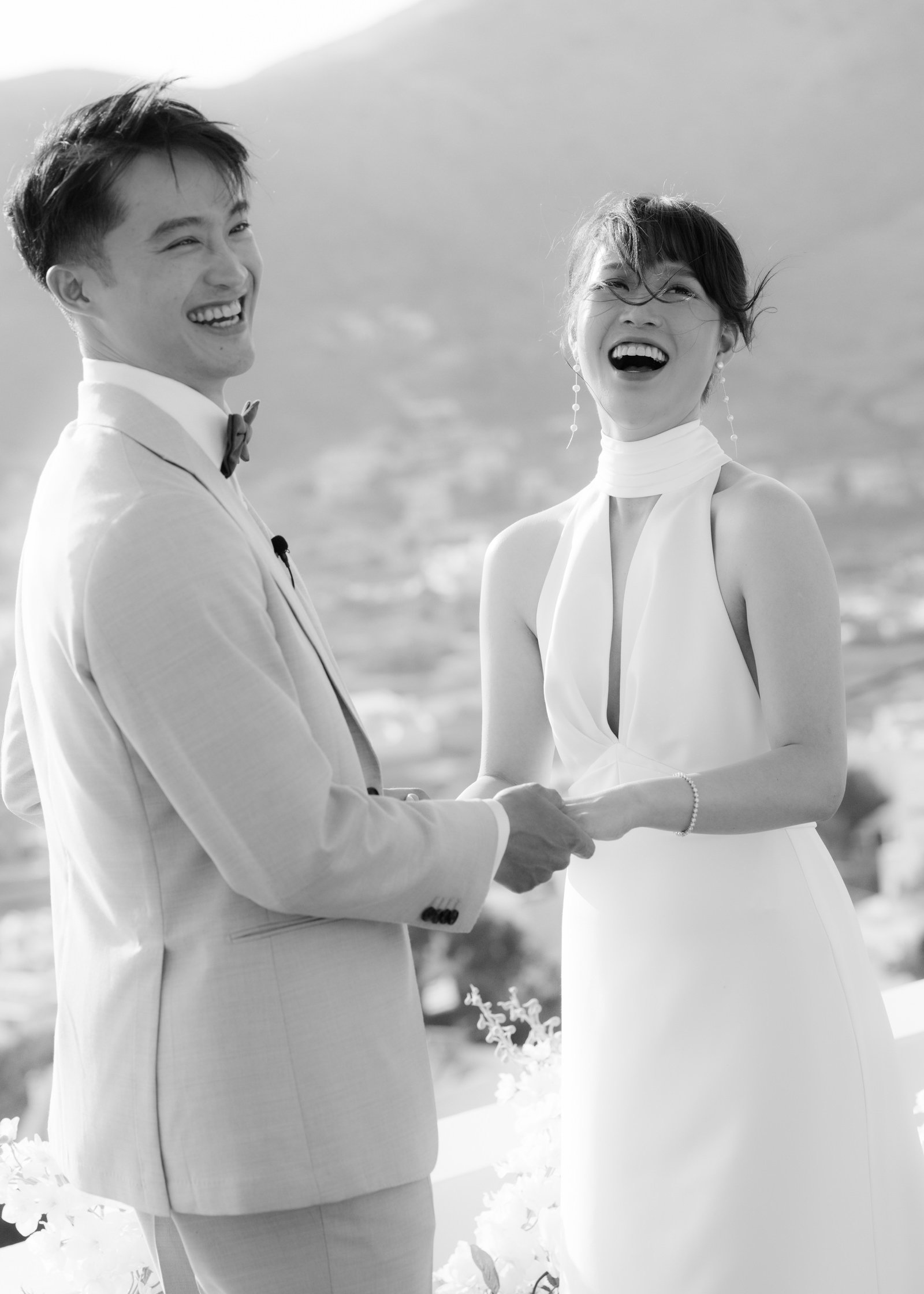 A black and white image of a laughing couple holding hand during a wedding ceremony outside.