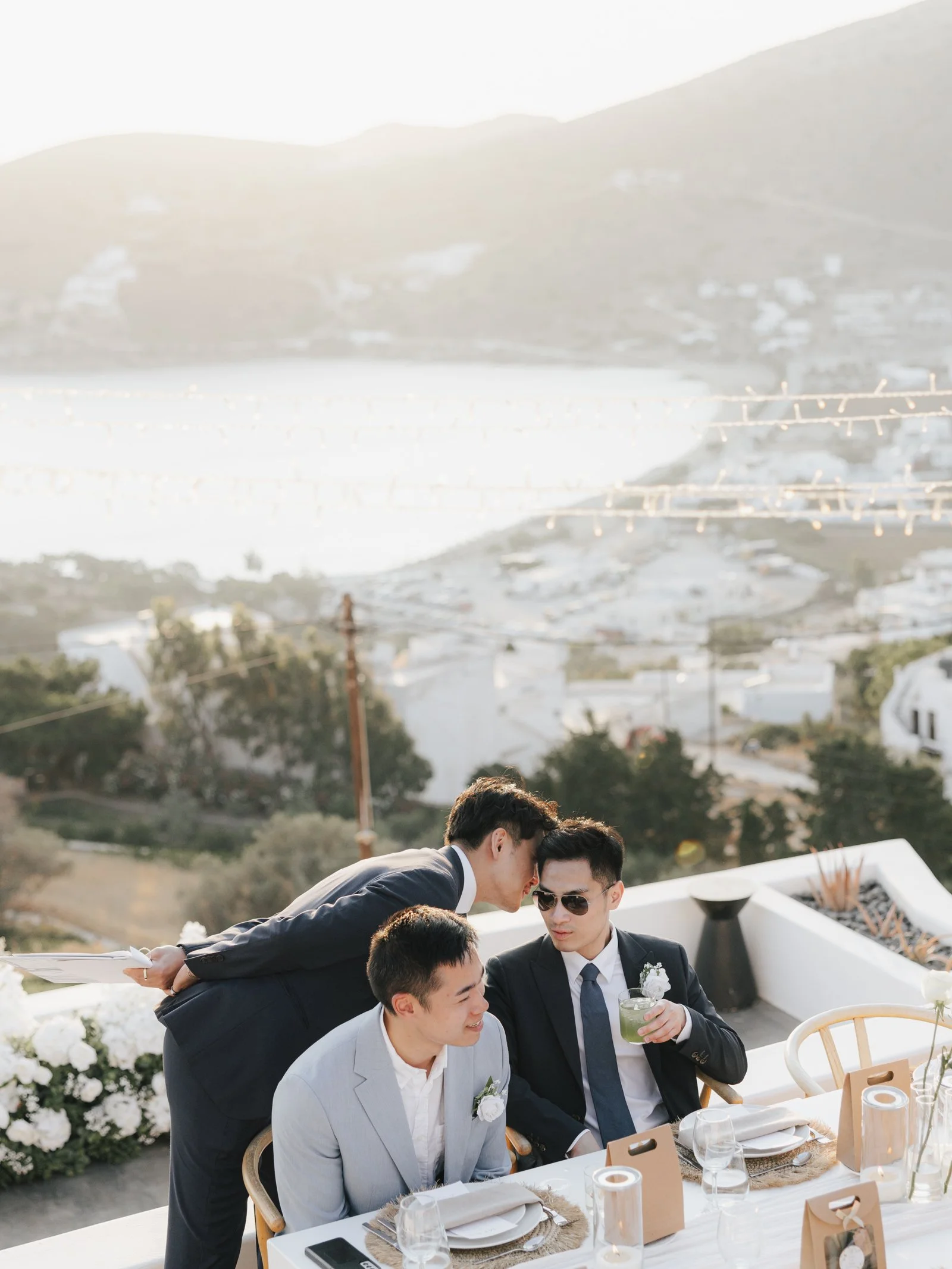 An outdoor scene of 3 men in suits at a dinner table. One man whispering into the other ones ear.