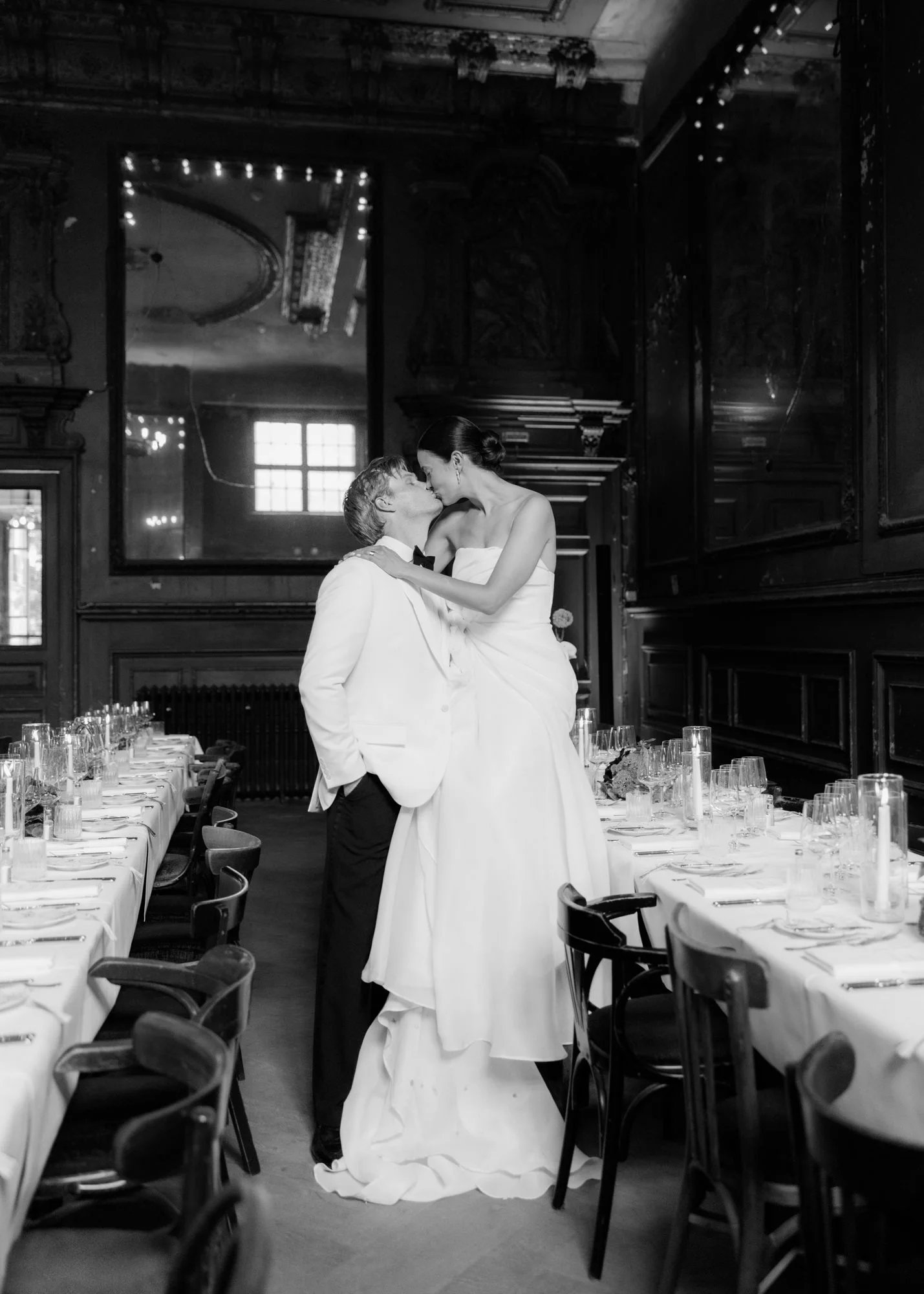 Black and white image of a bride and groom kissing in the middle of beautifully decorated dinner tables at Clärchens Ballhaus Berlin