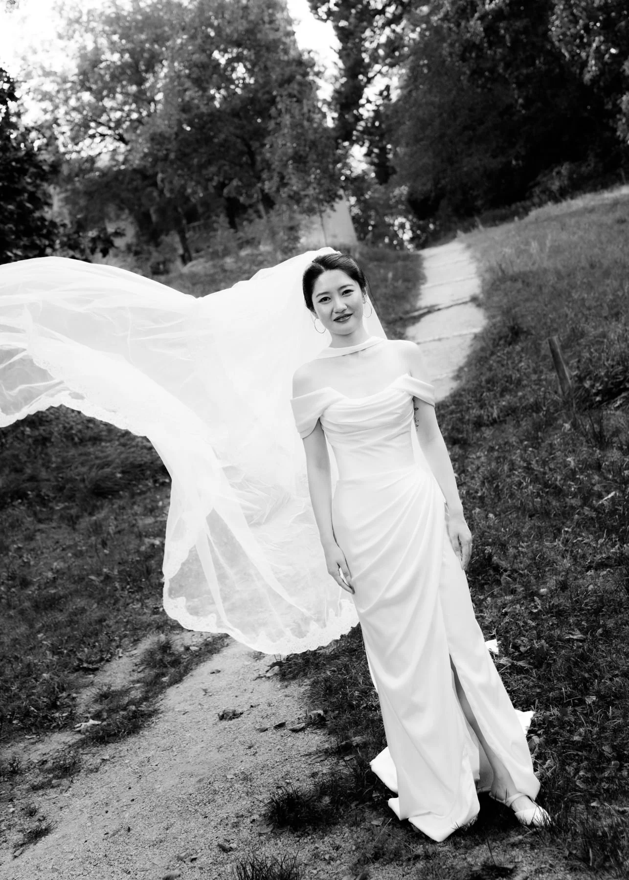 A woman in a wedding dress standing outdoors on a dirt path, holding her veil that flows behind her, surrounded by trees.