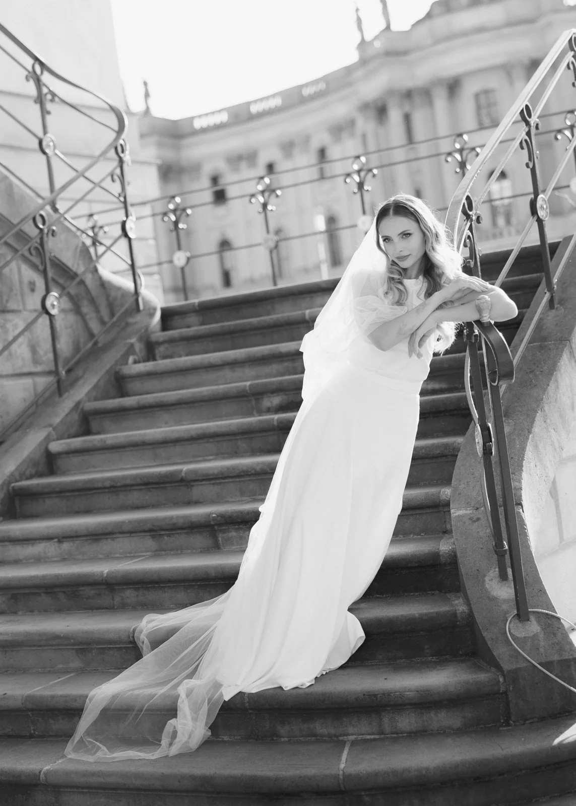Bride in an elegant gown and veil posing on a curved staircase in Berlin, with classical architecture in the background, captured in soft black and white.