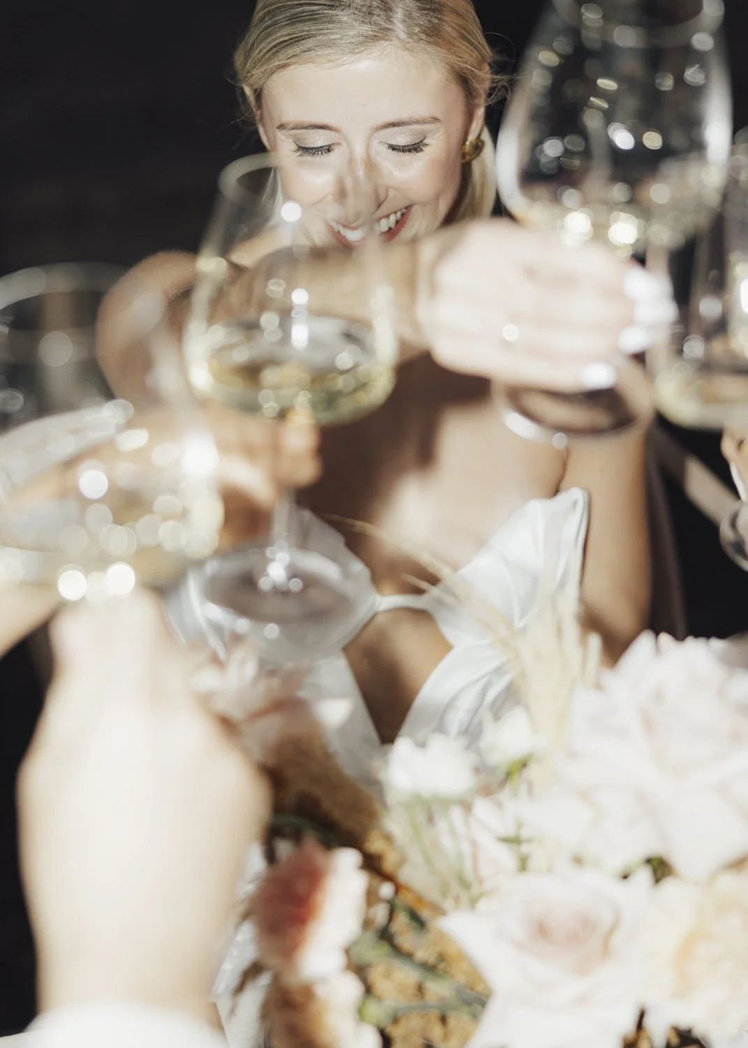 A joyful bride surrounded by guests raising glasses in a toast, her eyes closed with a radiant smile, holding a bouquet of pastel flowers.