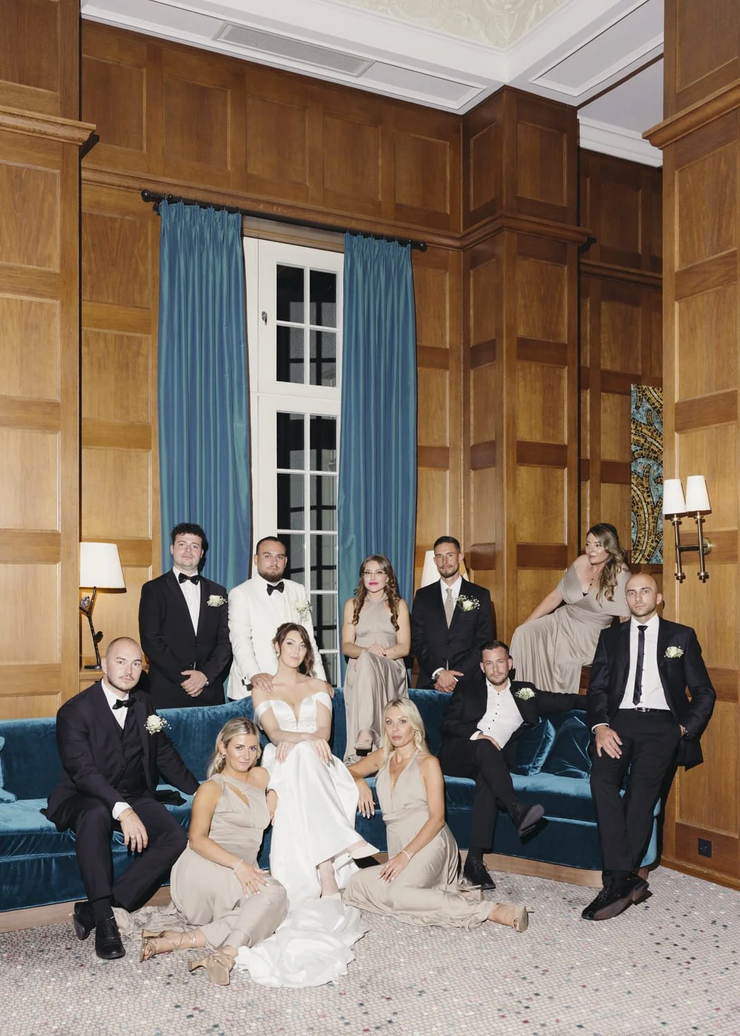 Formal wedding party portrait indoors with the bride and groom seated in the center, surrounded by bridesmaids in taupe dresses and groomsmen in black and white tuxedos. The scene is set in an elegant room with wood-paneled walls, teal velvet sofas,