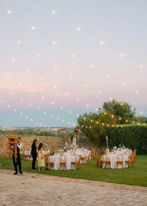 Outdoor event setup with round tables and chairs, string lights overhead, a tree, stone wall, and grassy area at sunset or dusk.
