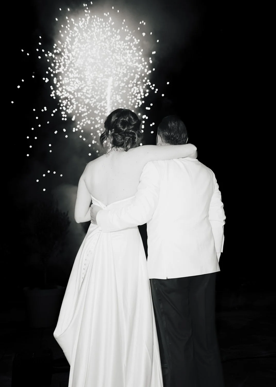 Bride and groom embracing while watching a fireworks display at night, captured in timeless black and white.