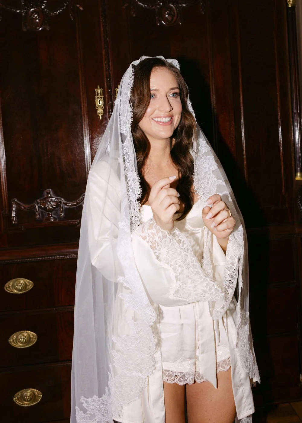 A woman in a white satin robe and lace veil, smiling and pointing her finger, standing in front of a dark wooden cabinet.