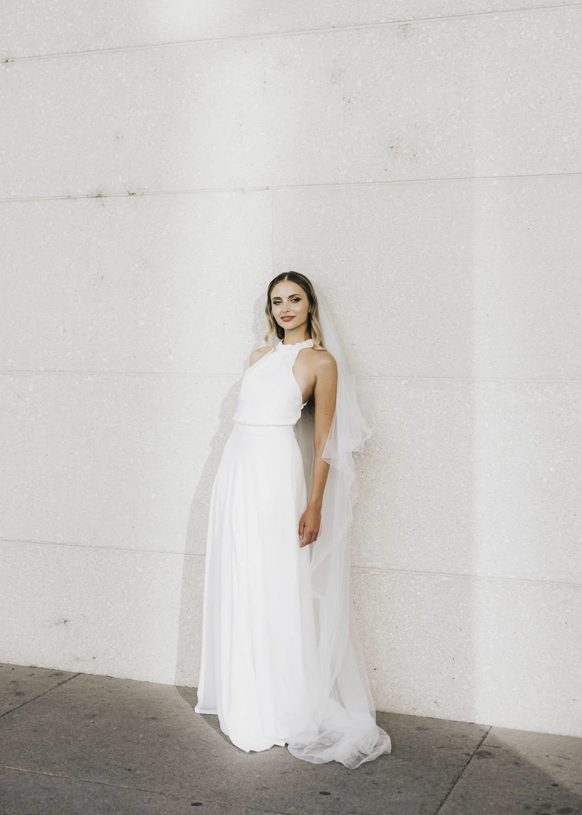 A bride in an elegant and modern dress leaning against a bright wall in Berlin in an editorial pose looking directly at the camera.