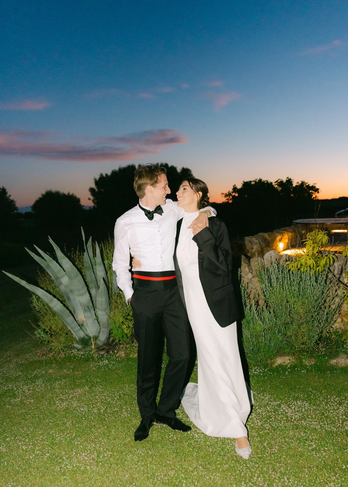 A couple dressed in formal attire, smiling and looking at each other, standing outdoors during sunset. The man is wearing a tuxedo with a bow tie, and the woman is in a white dress with a black jacket. There are lush plants and trees in the background.