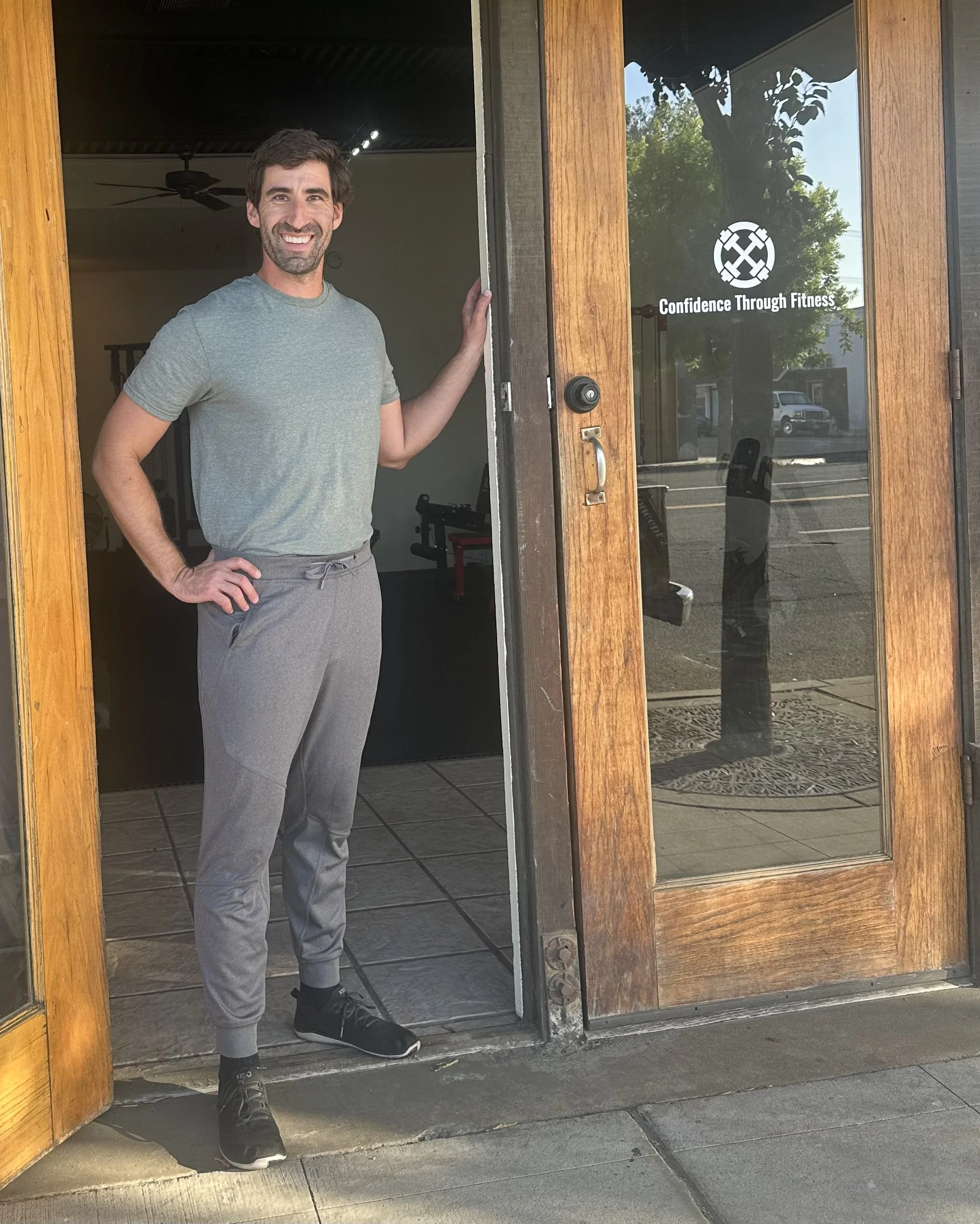 A man with dark hair and a beard standing in a doorway, smiling, wearing a grey T-shirt and grey sweatpants, inside a fitness studio with a ceiling fan, behind him a fitness equipment, with a sign on the glass door reading "Confidence Through Fitness."