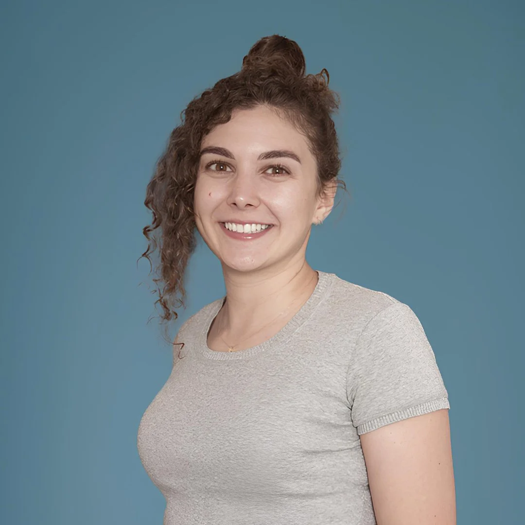 A young woman with curly hair in a high bun, smiling, wearing a light gray t-shirt against a solid blue background.