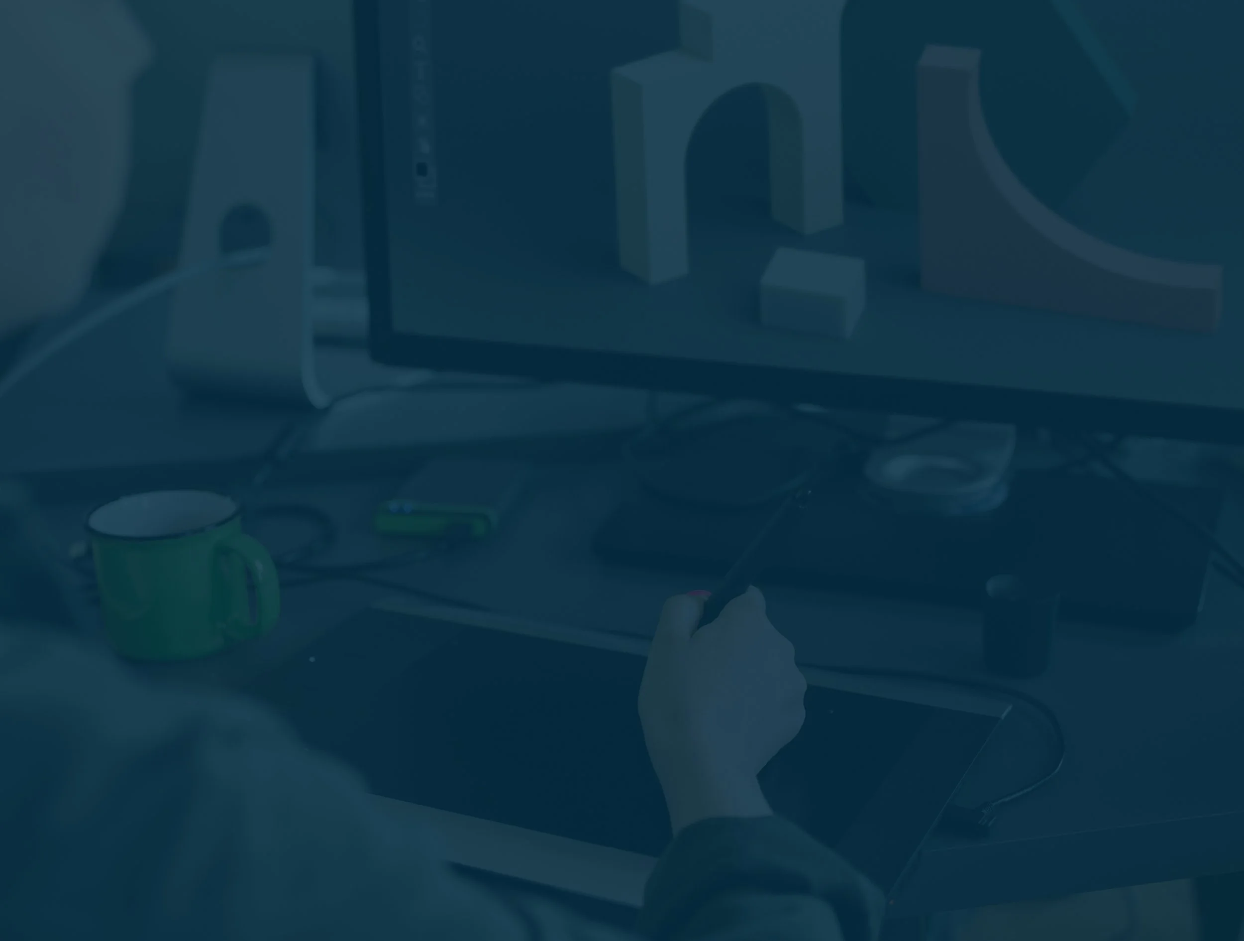 Person using a stylus on a graphics tablet exploring digital design, with a desktop monitor displaying 3D geometric shapes in the background, cluttered with office supplies.