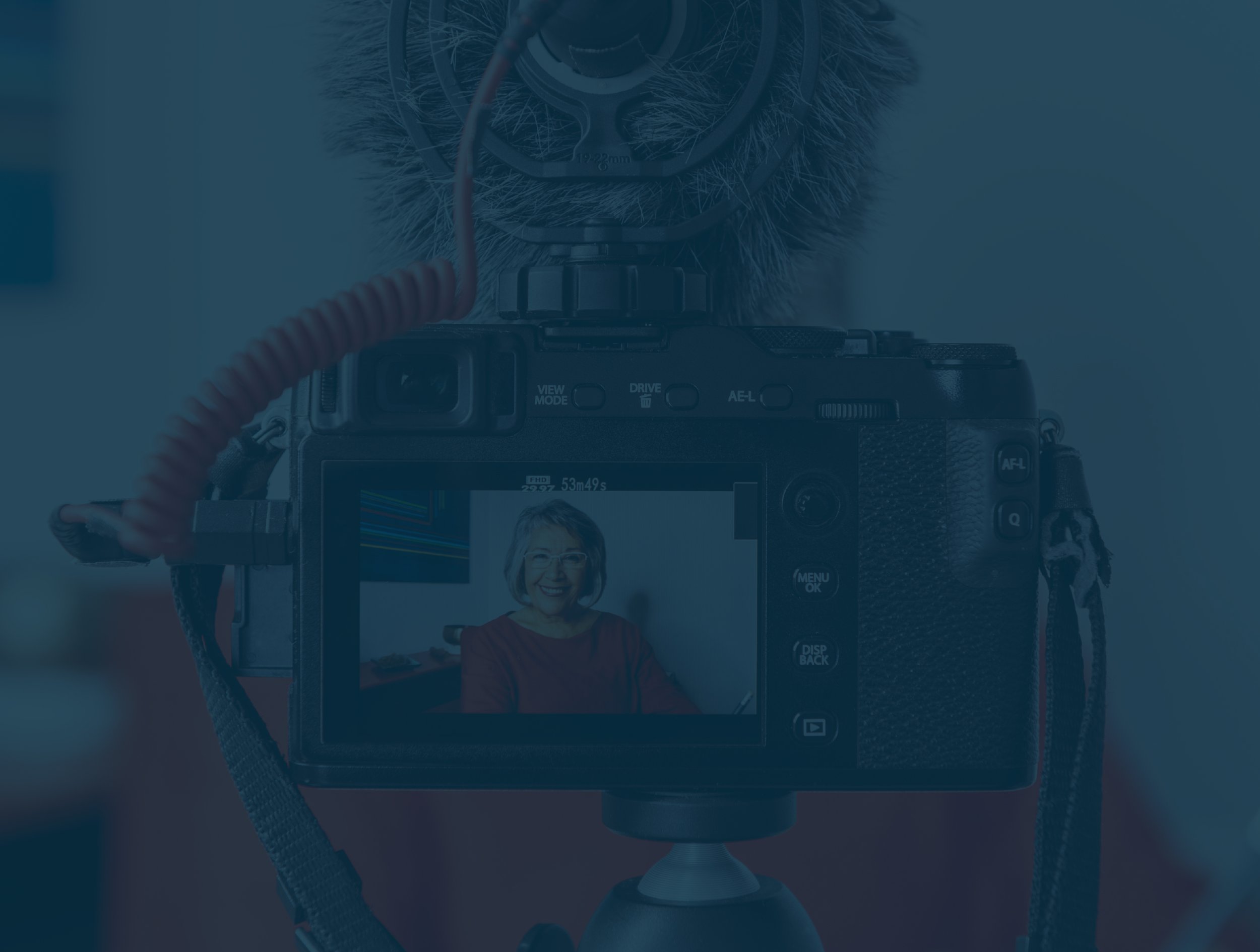A digital camera on a tripod capturing an image of a smiling woman with glasses, wearing a red top, in an indoor setting.
