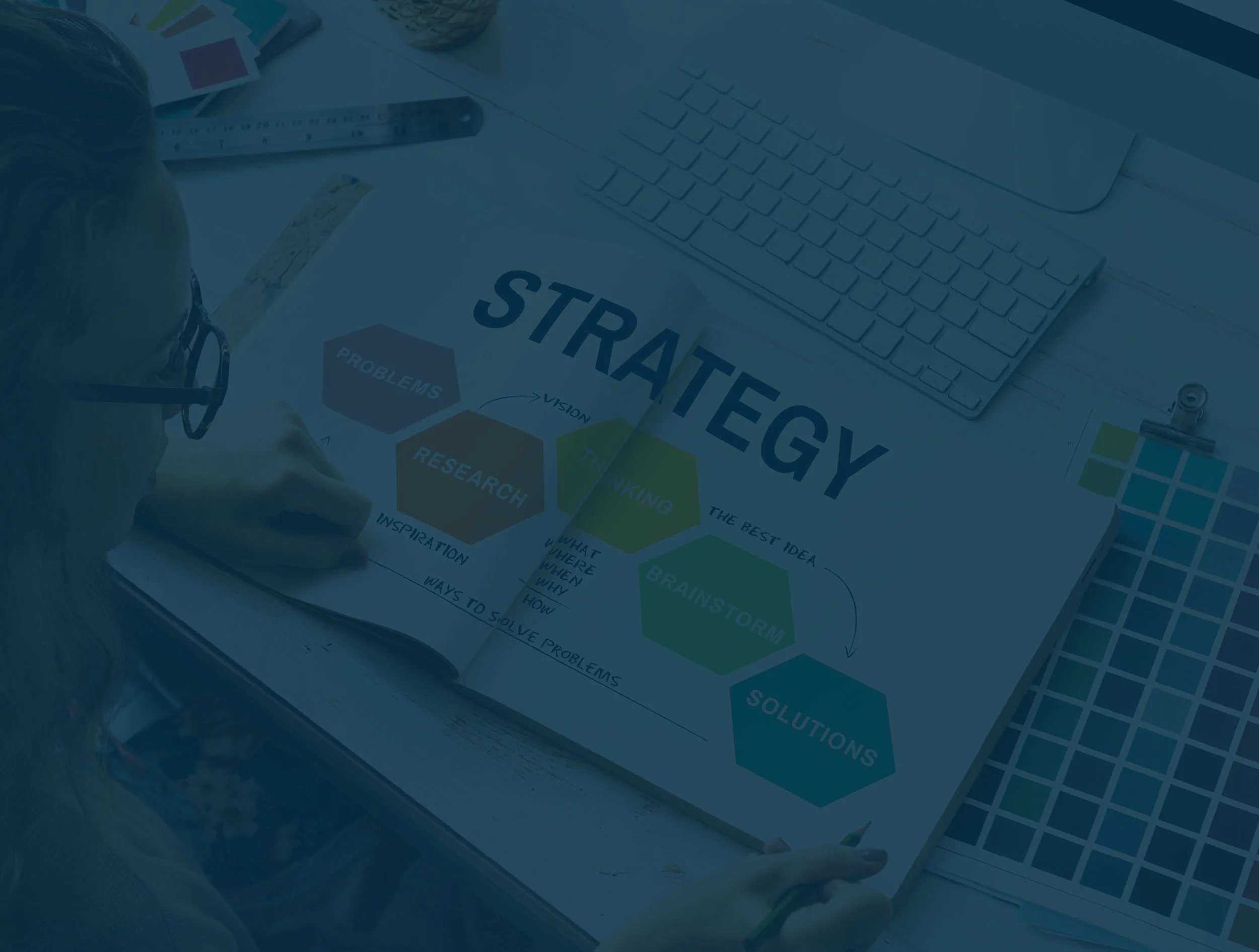 A person pointing to a document titled 'Strategy' on a desk, surrounded by a keyboard, ruler, color swatches, and stationery.