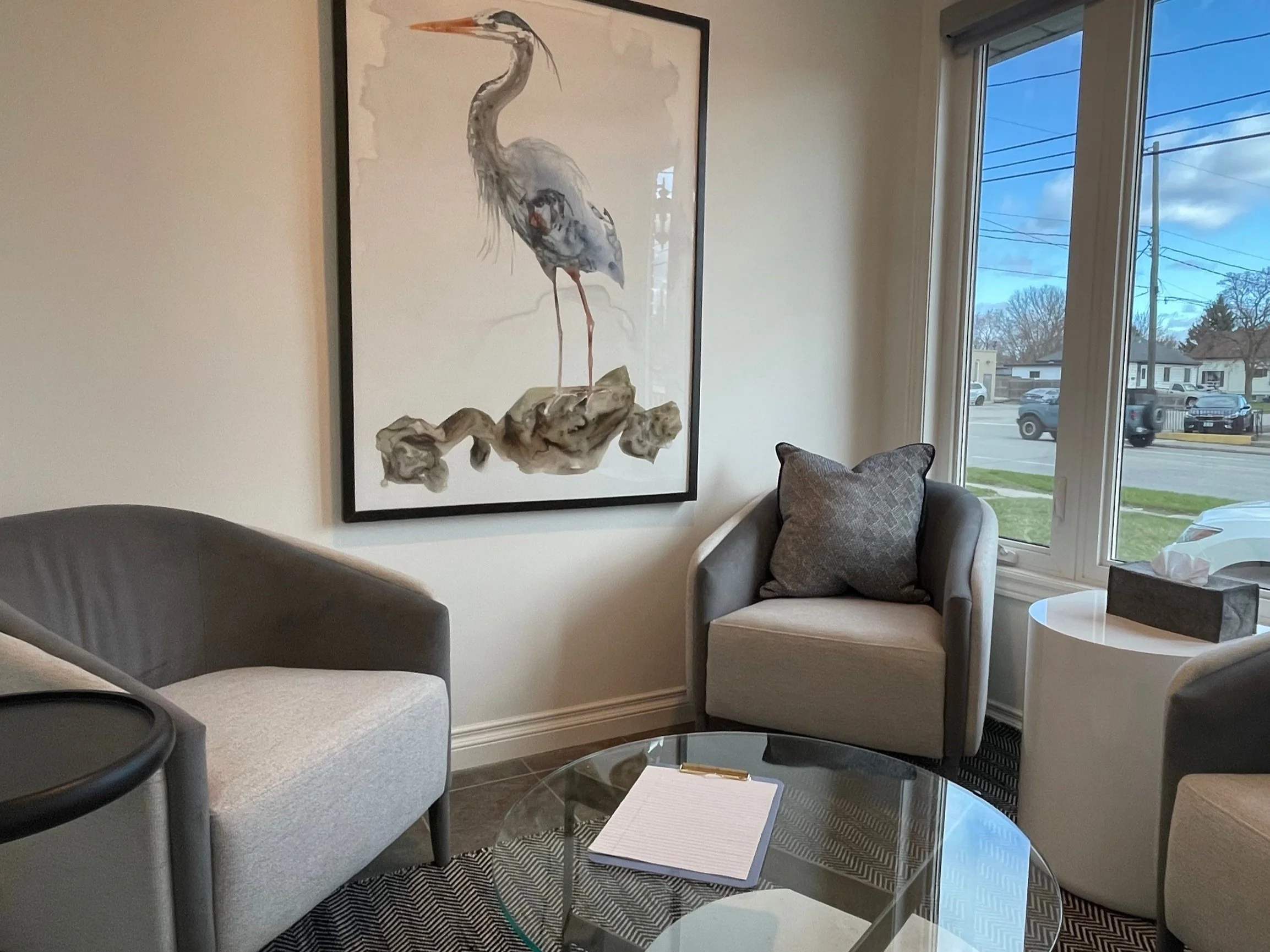 A cozy waiting area with gray and beige chairs, a round glass coffee table with a clipboard, a window with a view of the street, and a large framed painting of a heron on the wall.