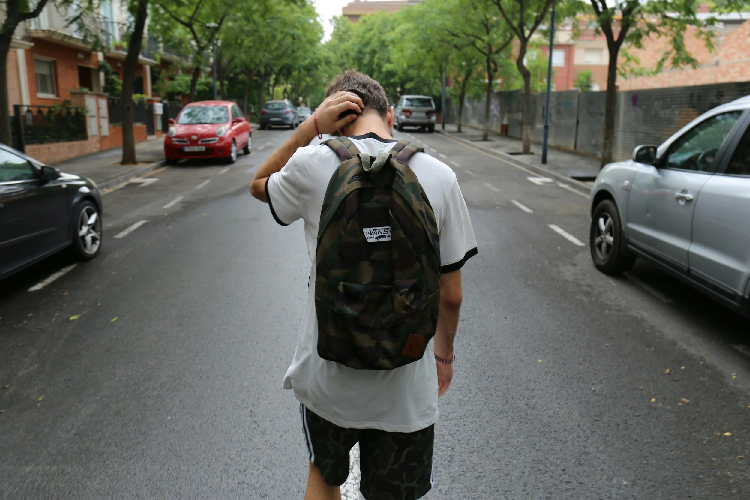 Young man with a camouflage backpack walking on a residential street with trees, parked cars, and brick buildings