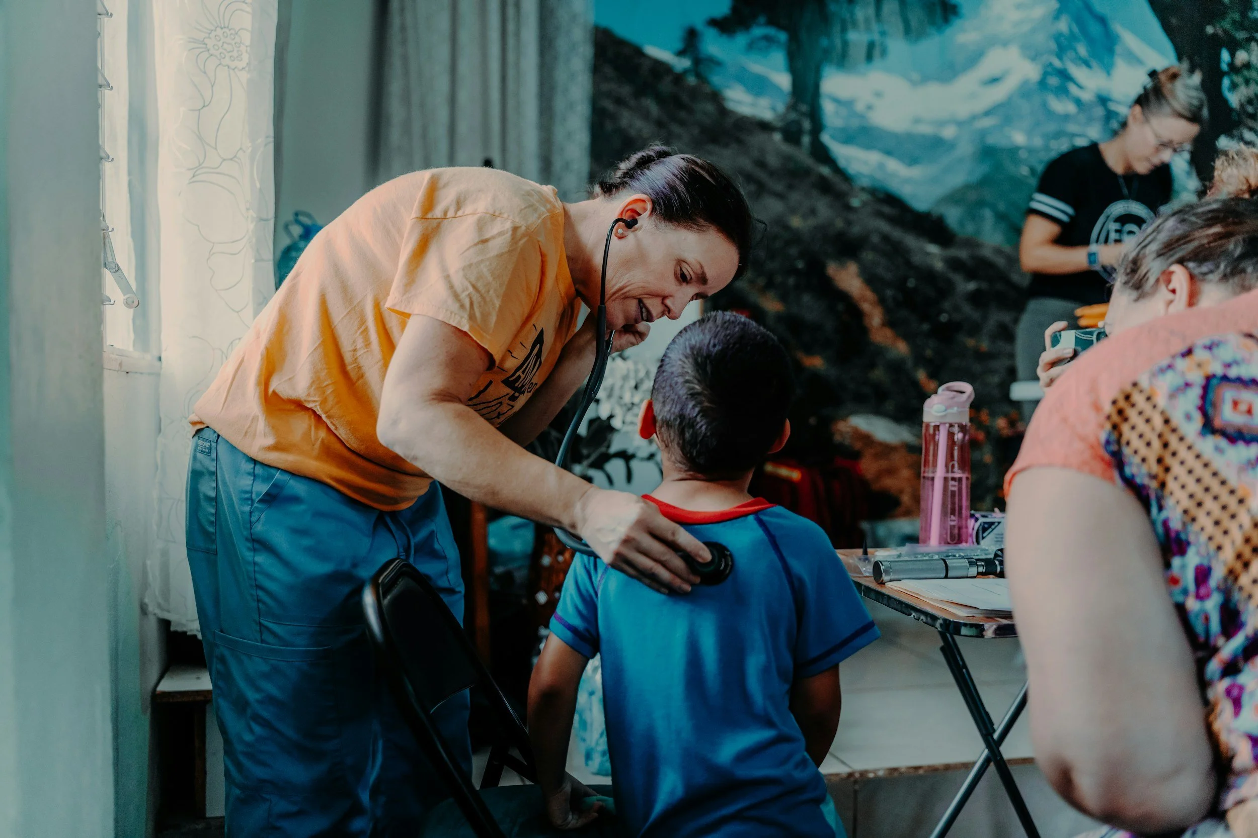 A healthcare professional using a stethoscope to examine a young child's back in a clinic setting with a mountainous mural in the background.