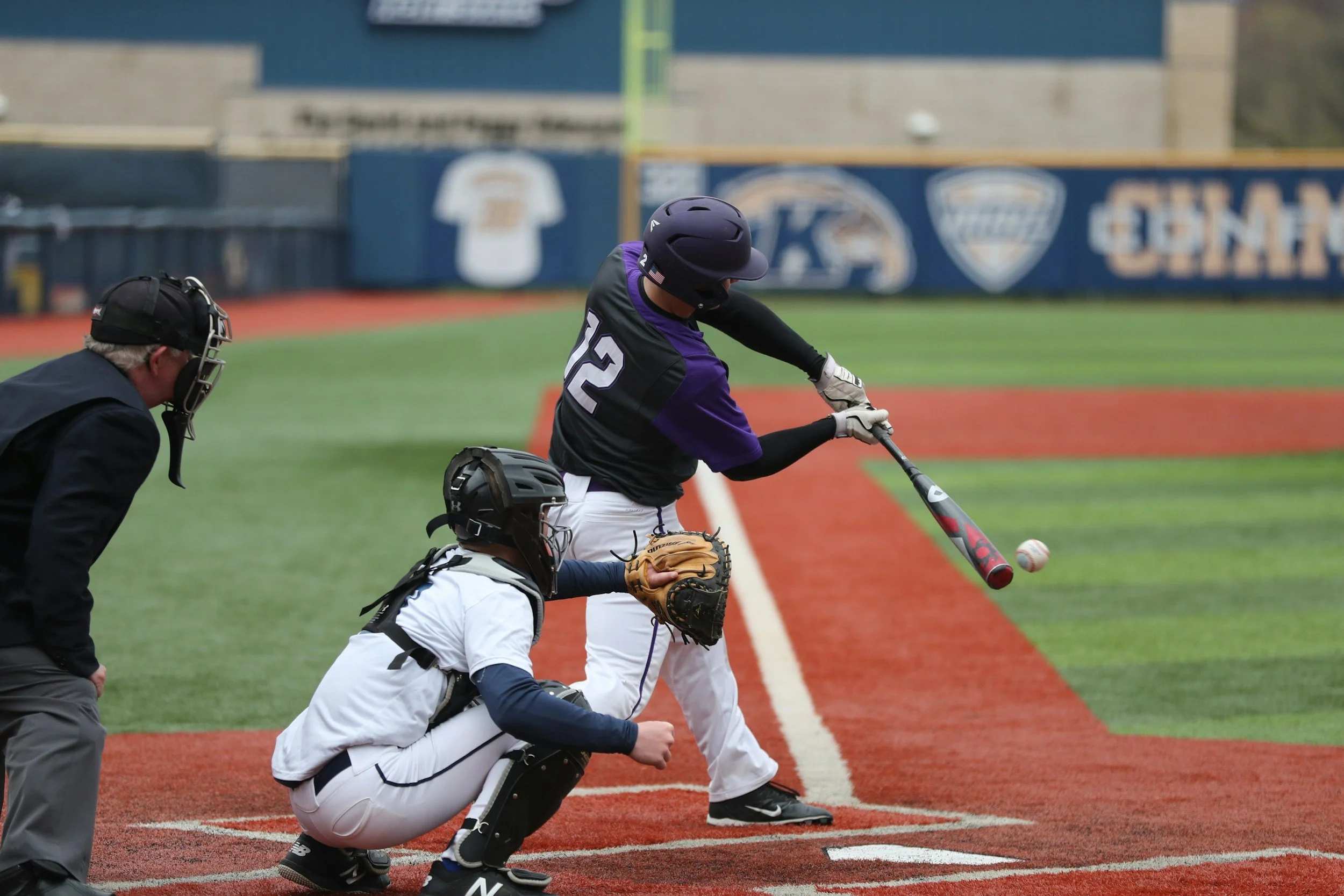 A baseball player swings a bat at a pitched ball during a game, with an umpire and catcher behind him on the field.