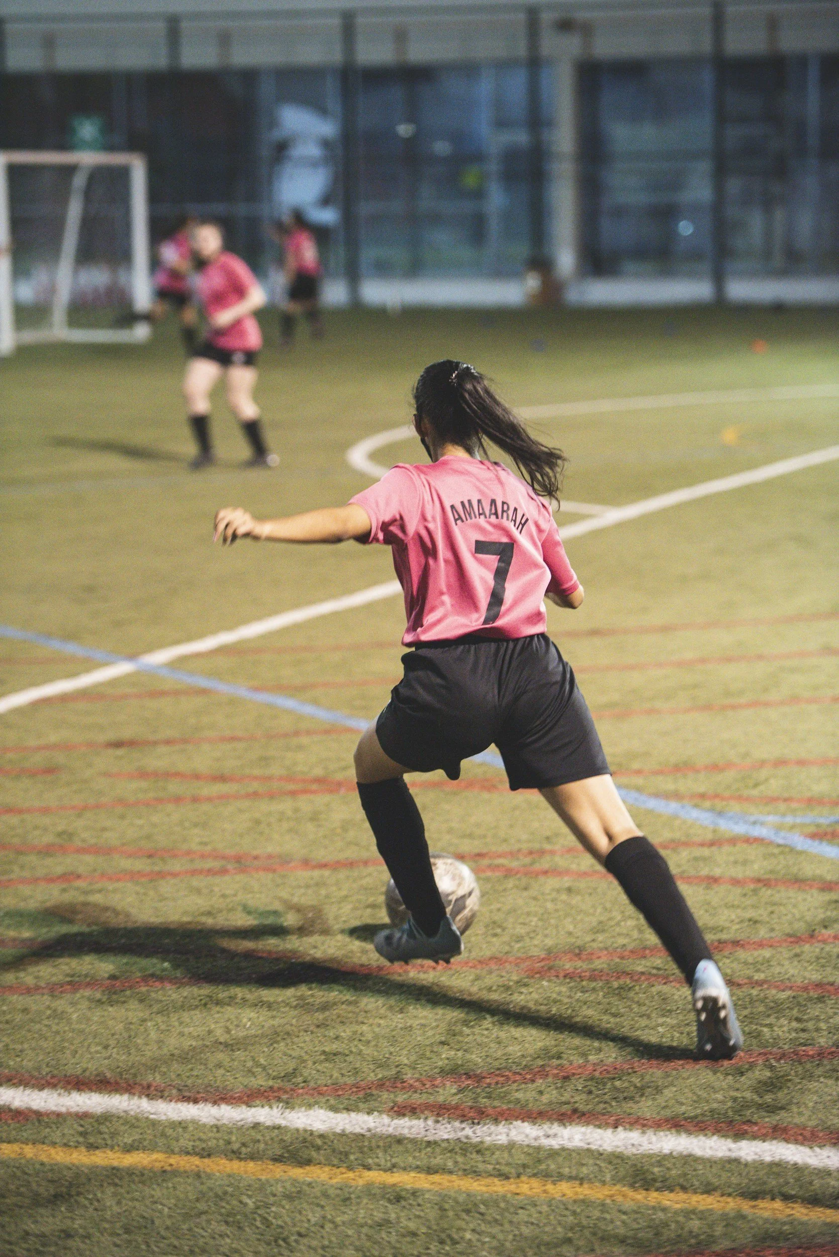 A female soccer player wearing a pink jersey with the name 'AMARAH' and the number 7, is in the process of kicking a soccer ball on an indoor soccer field. Other players are visible in the background.