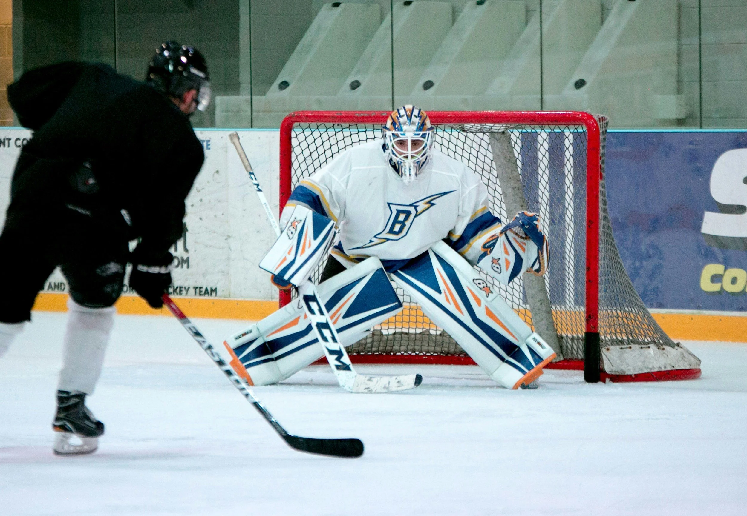 Hockey goalie in white and blue gear ready to block a shot, facing an opposing player in black on an ice rink.