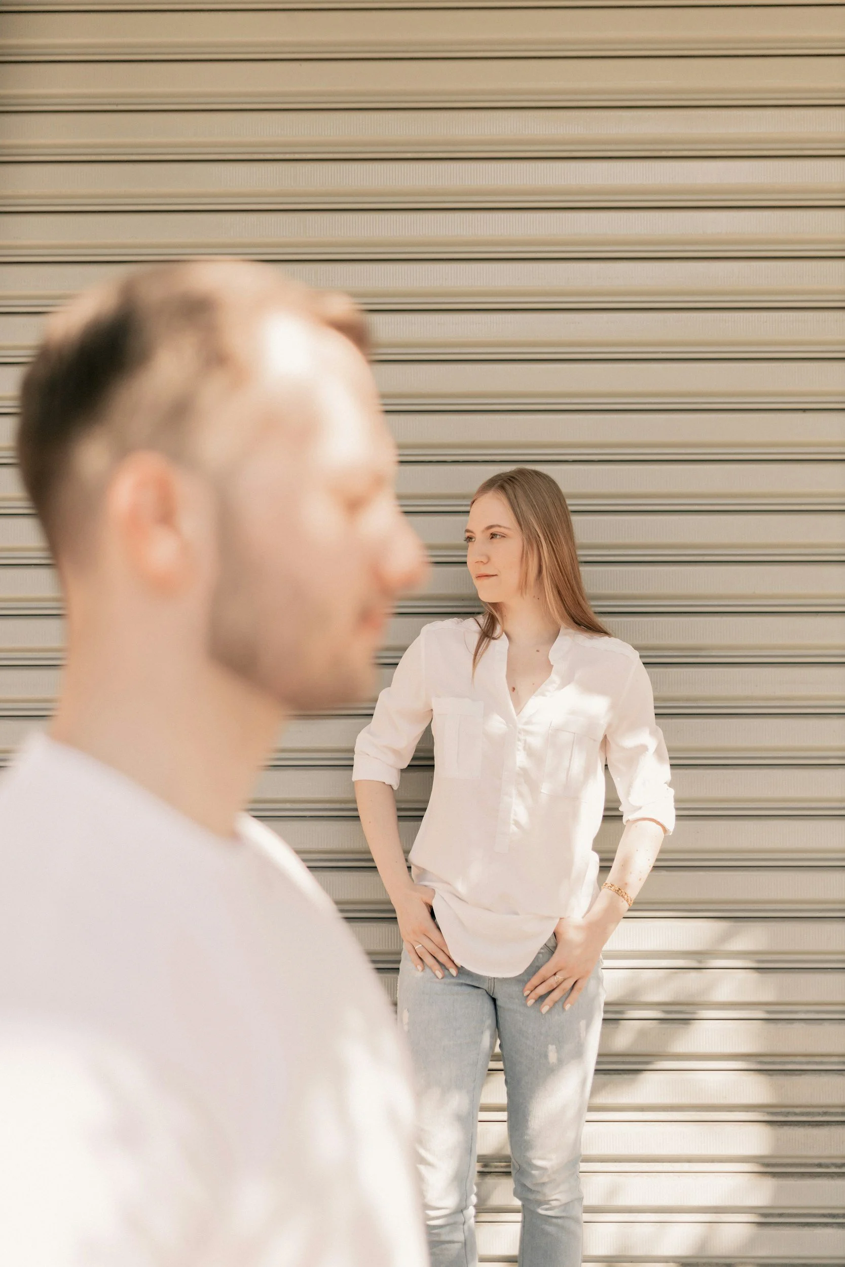 A woman in a white shirt and light gray pants standing against a metal roll-up door, looking at a blurred man in the foreground.