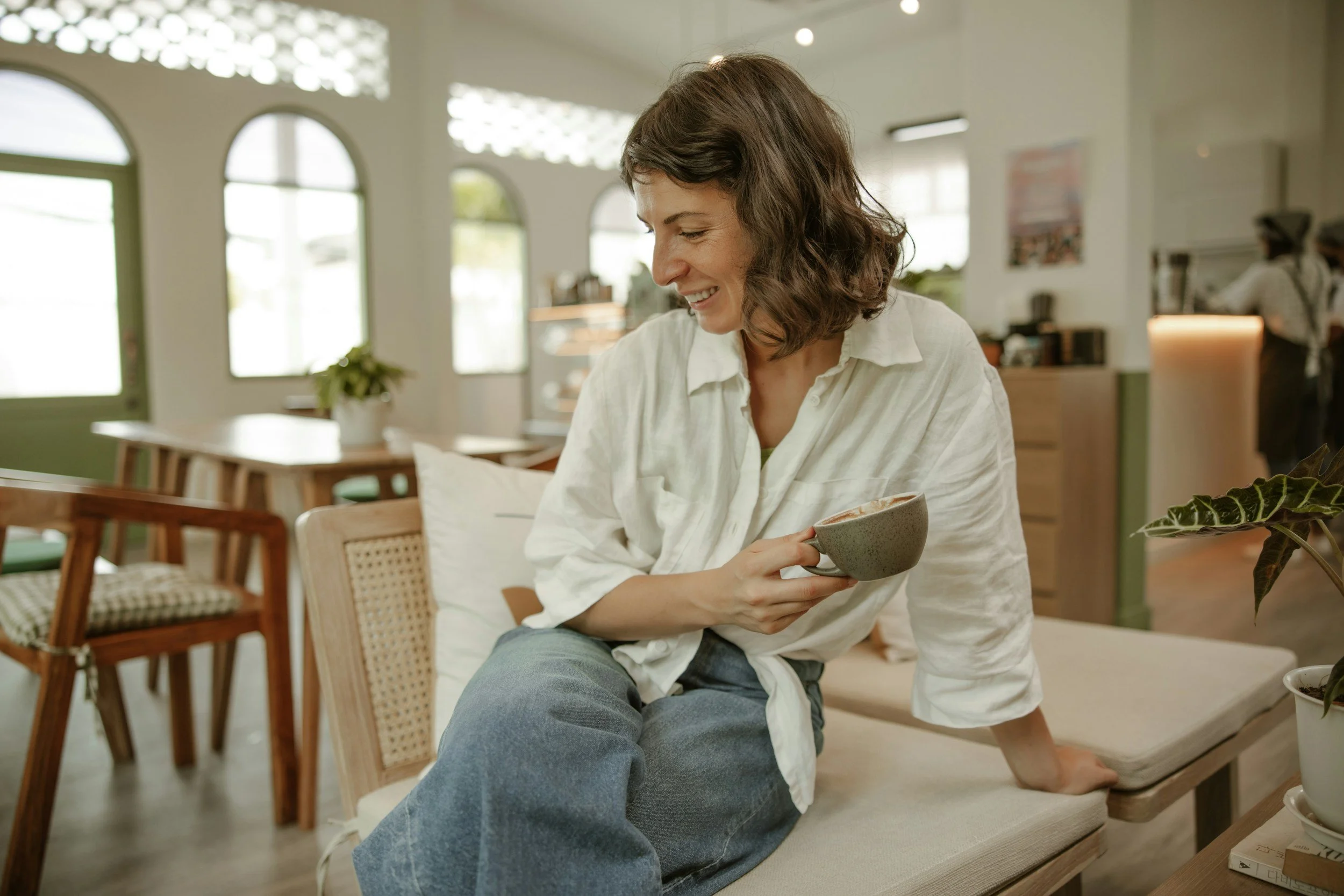 A woman with short brown hair and a white shirt sitting on a cushioned bench in a cozy cafe, smiling and holding a cup with a hot beverage, surrounded by indoor plants and wooden furniture.