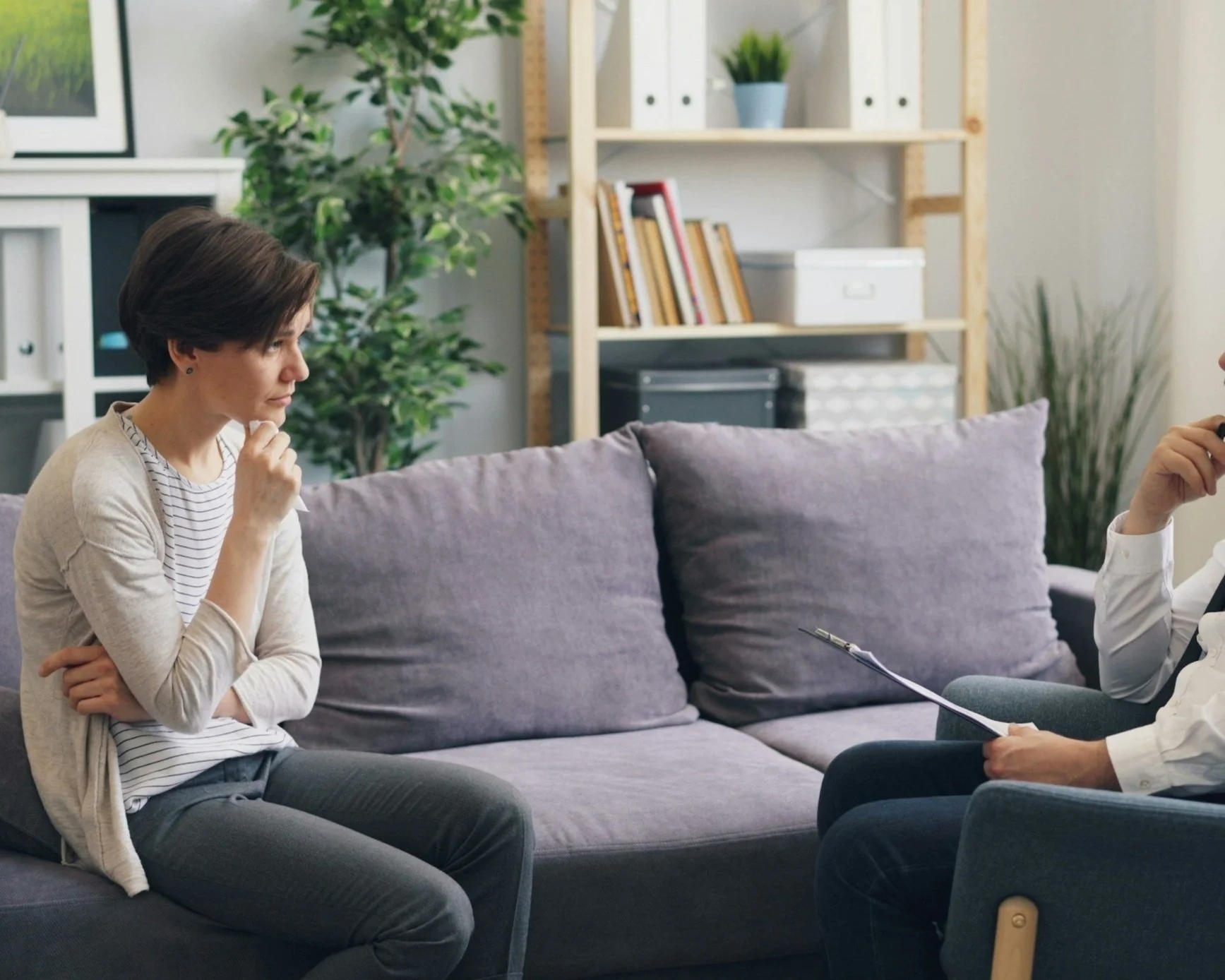 A woman attending a therapy session sitting on a couch, with a therapist holding a clipboard across from her. The room is decorated with bookshelves, plants, and framed art.