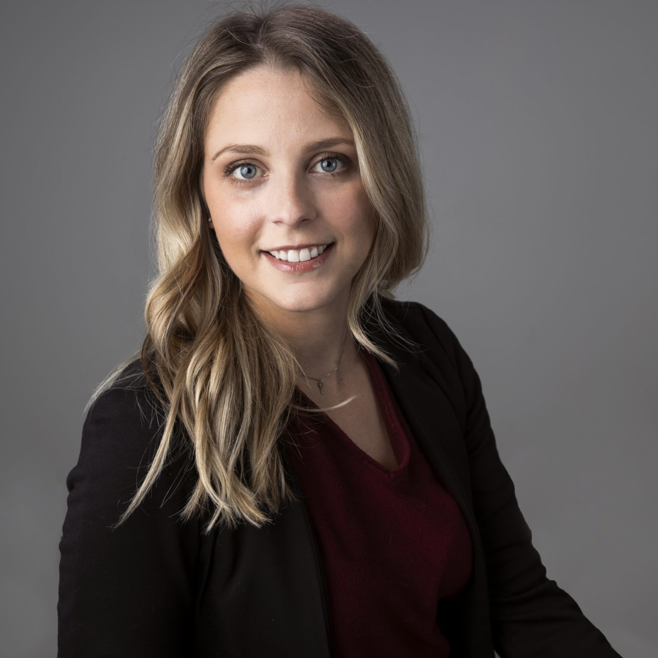 Portrait of a young woman with long wavy blonde hair, blue eyes, and a friendly smile, wearing a black jacket and a dark red top, against a gray background.