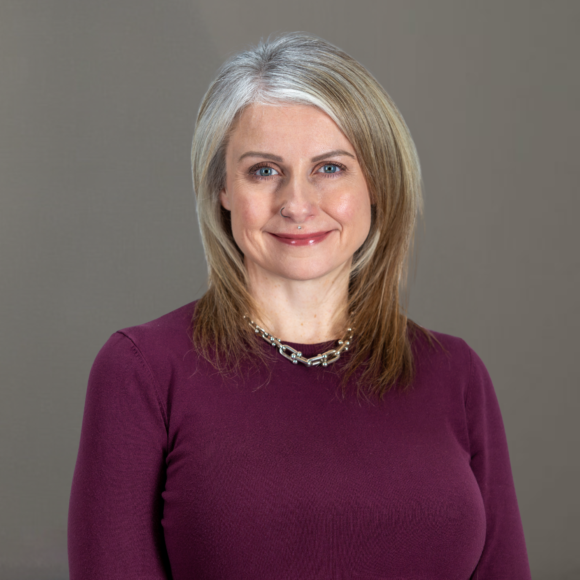 A smiling woman with shoulder-length blonde hair, wearing a purple top and a silver necklace, poses against a plain gray background.