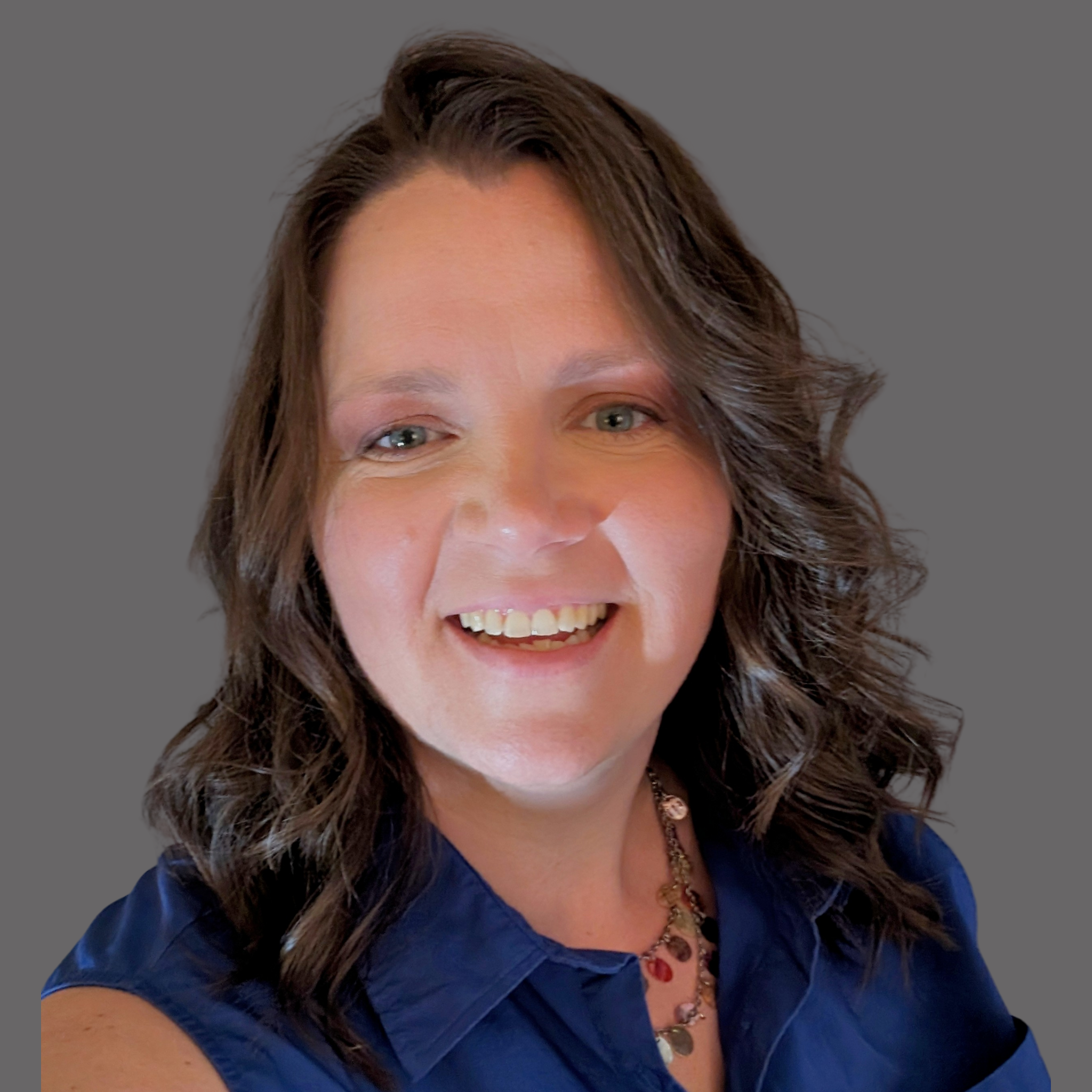 A woman with shoulder-length brown curly hair smiling, wearing a blue top and a necklace, against a gray background.