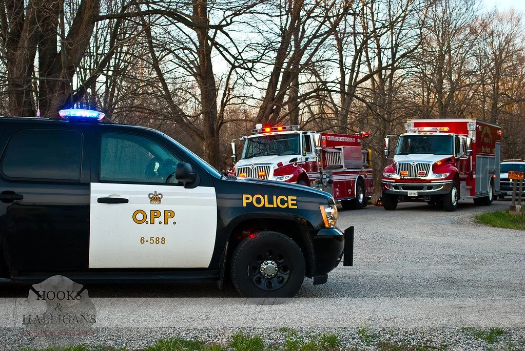 A police SUV with blue lights on, and two fire trucks parked behind it on a gravel road with leafless trees in the background.