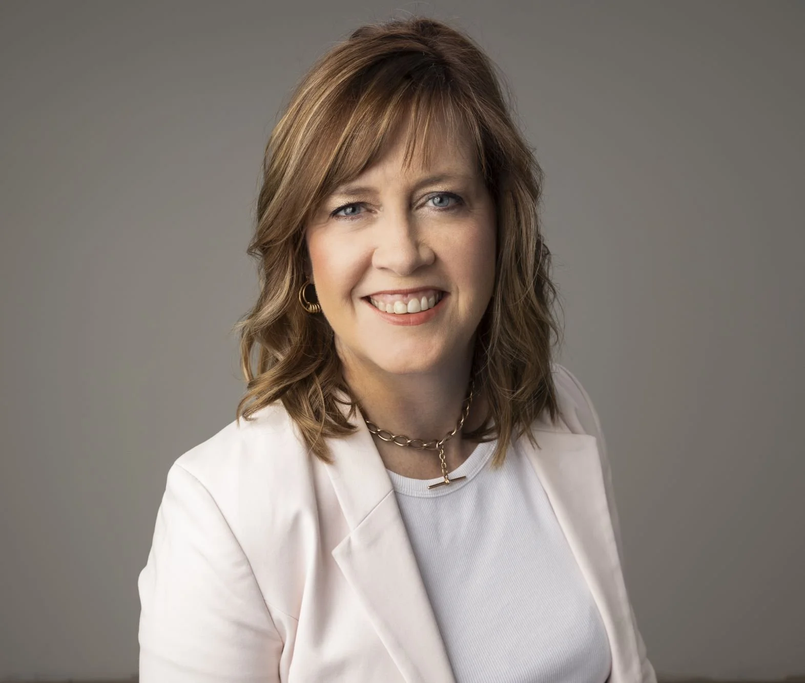 A woman with shoulder-length brown hair, blue eyes, and fair skin smiling, wearing a white blazer, white top, gold hoop earrings, a chain necklace, and a shimmery necklace, against a gray background.
