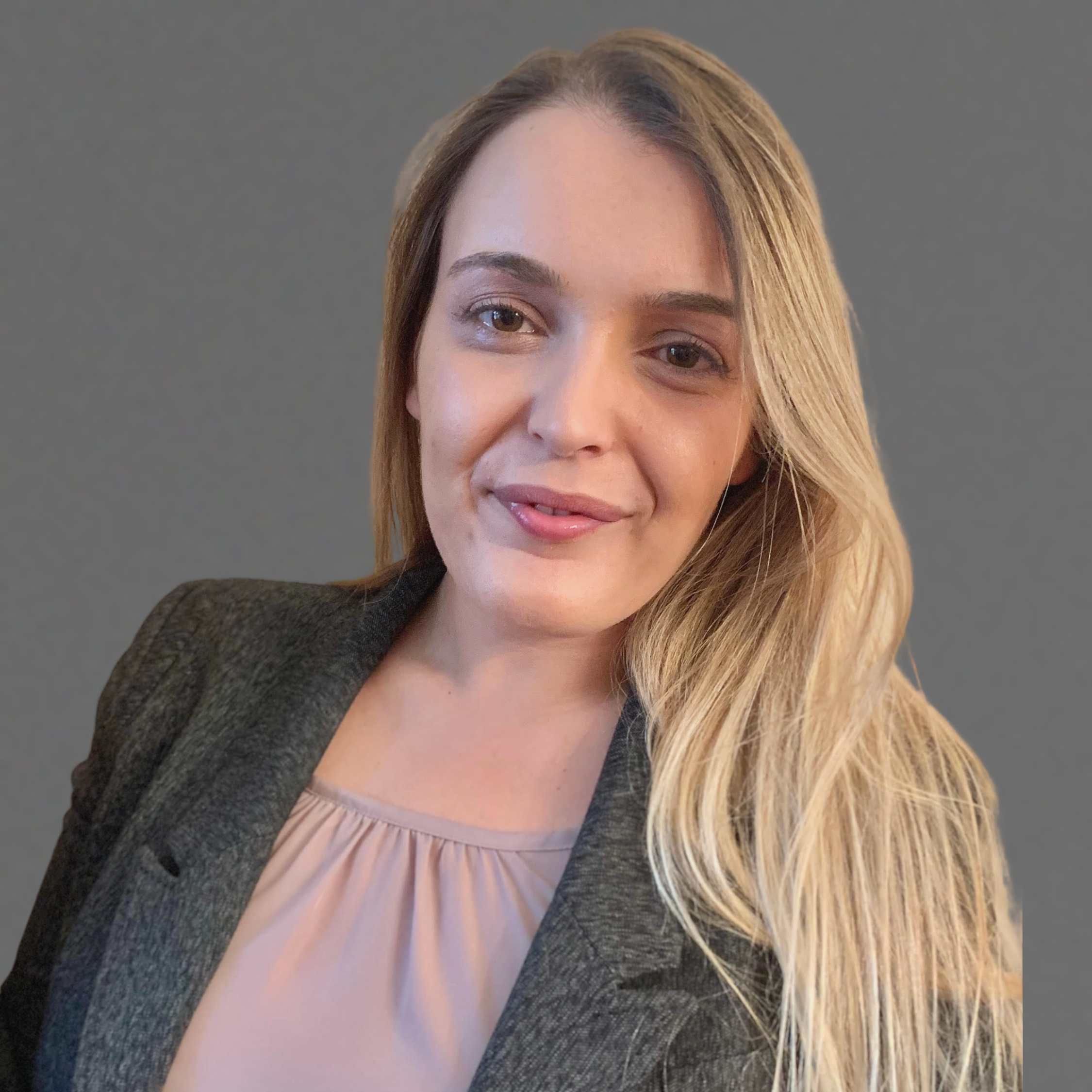A woman with long blonde hair wearing a grey blazer over a light pink blouse, smiling slightly, against a plain grey background.