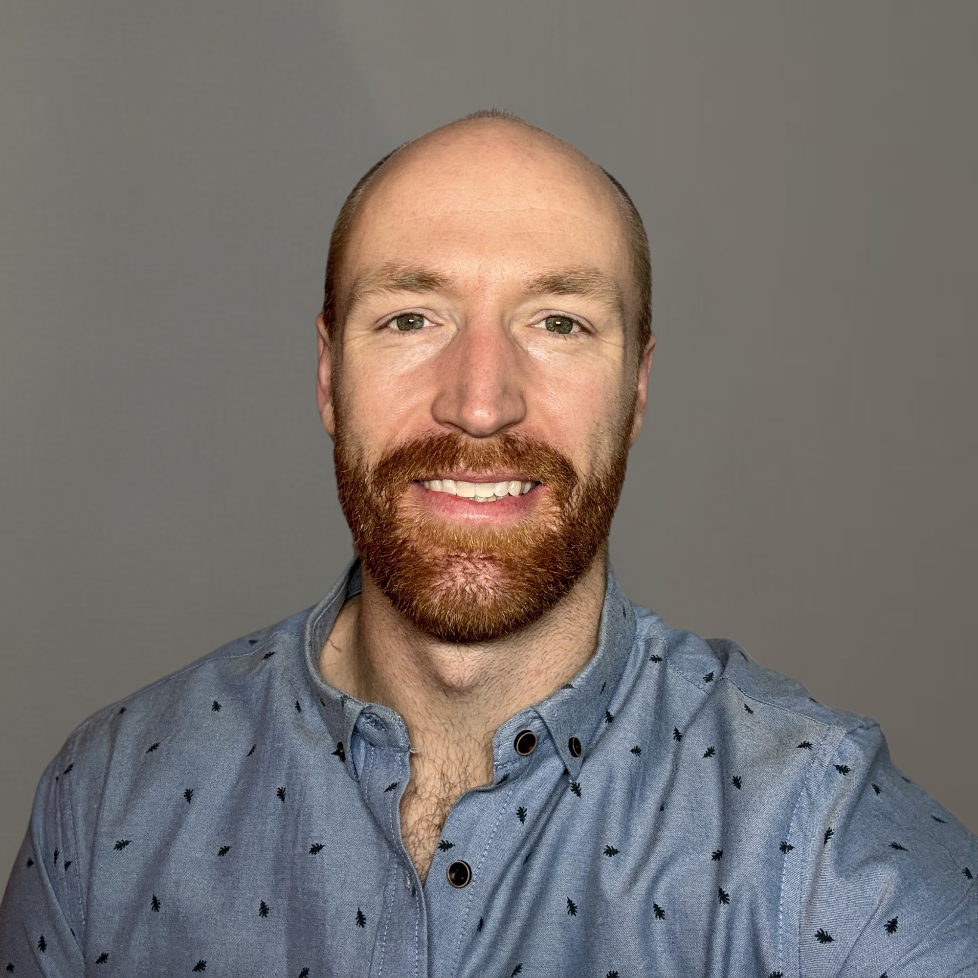 Portrait of a man with a beard and mustache, smiling, wearing a light blue button-up shirt with small patterns, against a plain gray background.