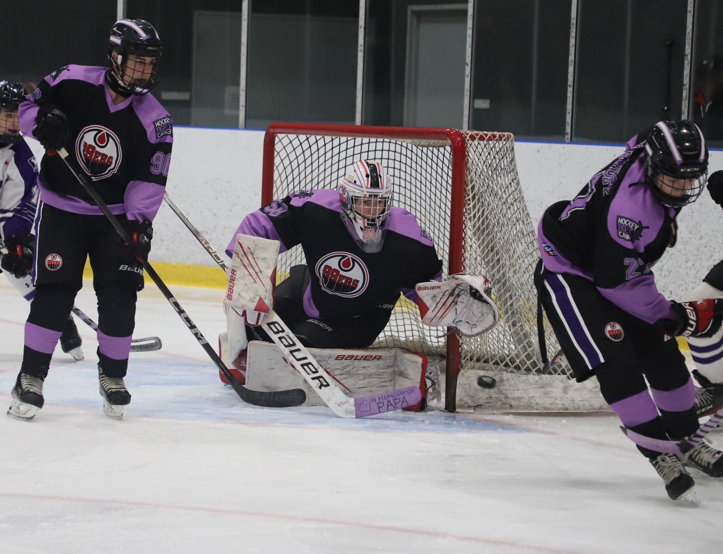 Ice hockey players in black and purple uniforms contest a game near the goal, with a goalie in a purple and black uniform crouched in front of the net.