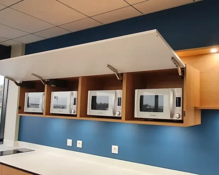 Meuble sur mesure pour salle de pause d’entreprise à Clermont-Ferrand, conçu par Oleksiak. Rangements intégrés avec portes relevables et emplacements pour fours micro-ondes, alliant praticité et design en bois clair et bleu profond.