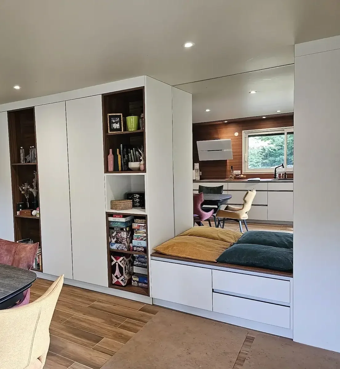 Intérieur d'une maison avec espace bibliothèque, coin lecture et cuisine ouverte, mobilier contemporain, sol en bois, fenêtre avec vue sur la nature.